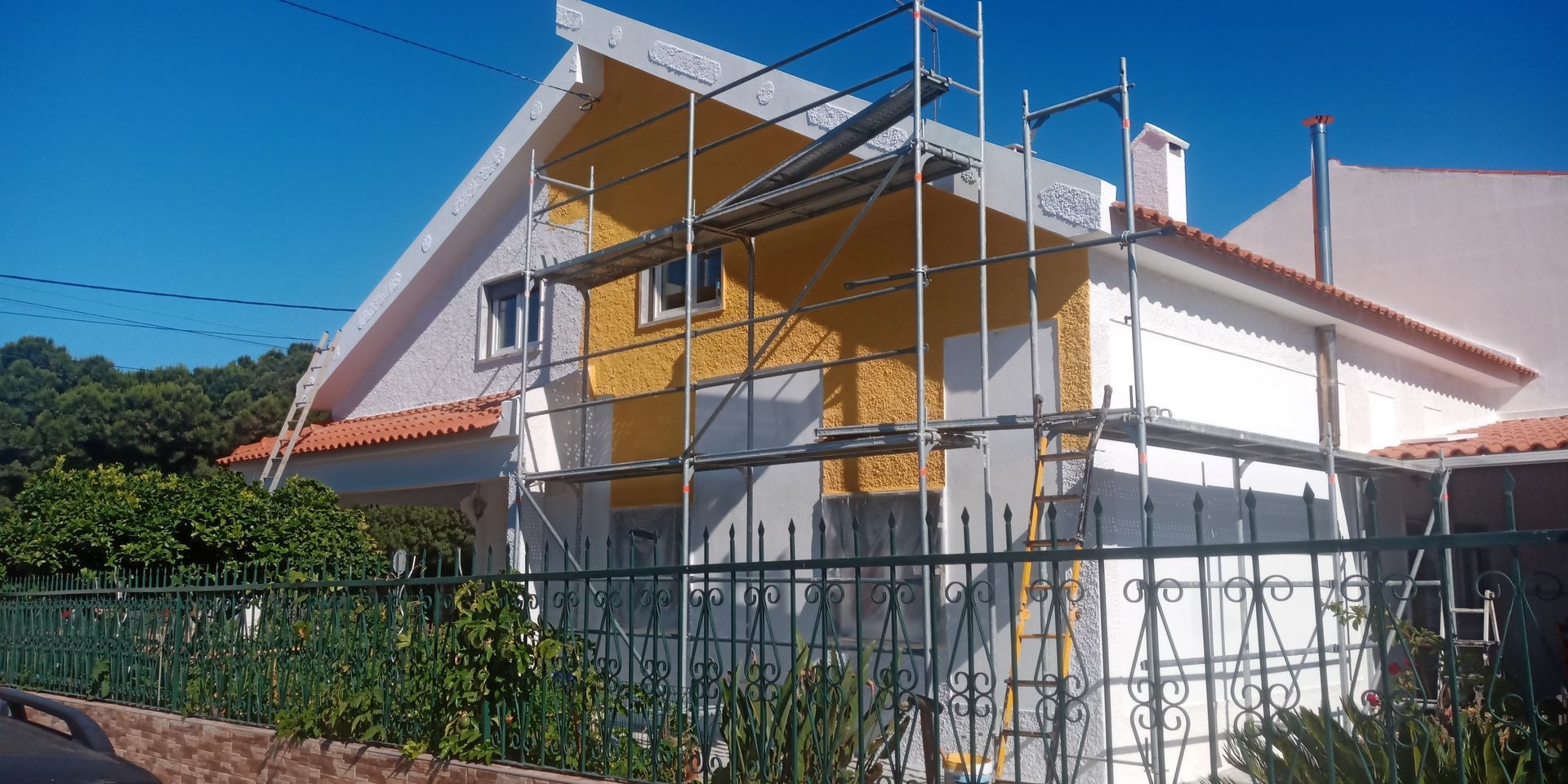 Pintura externa da casa, com andaimes no local. Casa amarela e branca sob um céu azul claro.