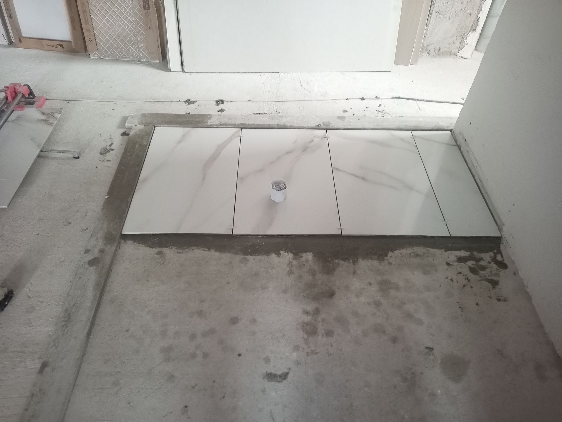 Piso de azulejo branco recém-instalado com ralo, rejunte e concreto inacabado ao redor em um cômodo.