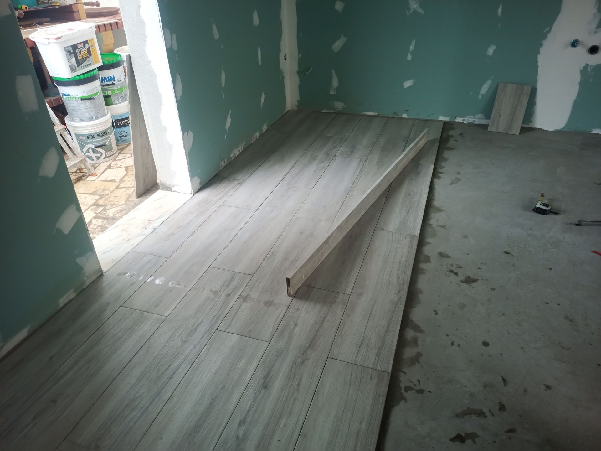 Azulejos cinza imitando madeira assentados sobre piso de concreto em um cômodo em construção. Um nível está apoiado sobre os azulejos.