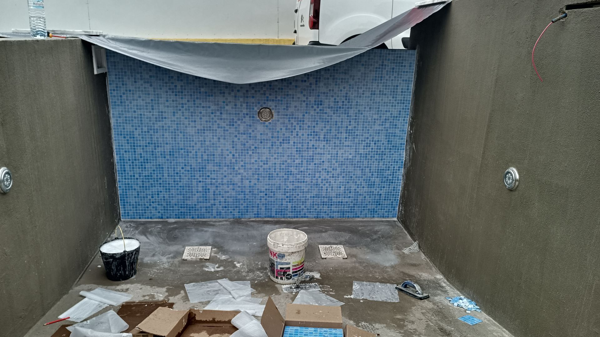 Construção de piscina: parede revestida com azulejos azuis, paredes de cimento cinza, baldes, caixas e entulho no chão.