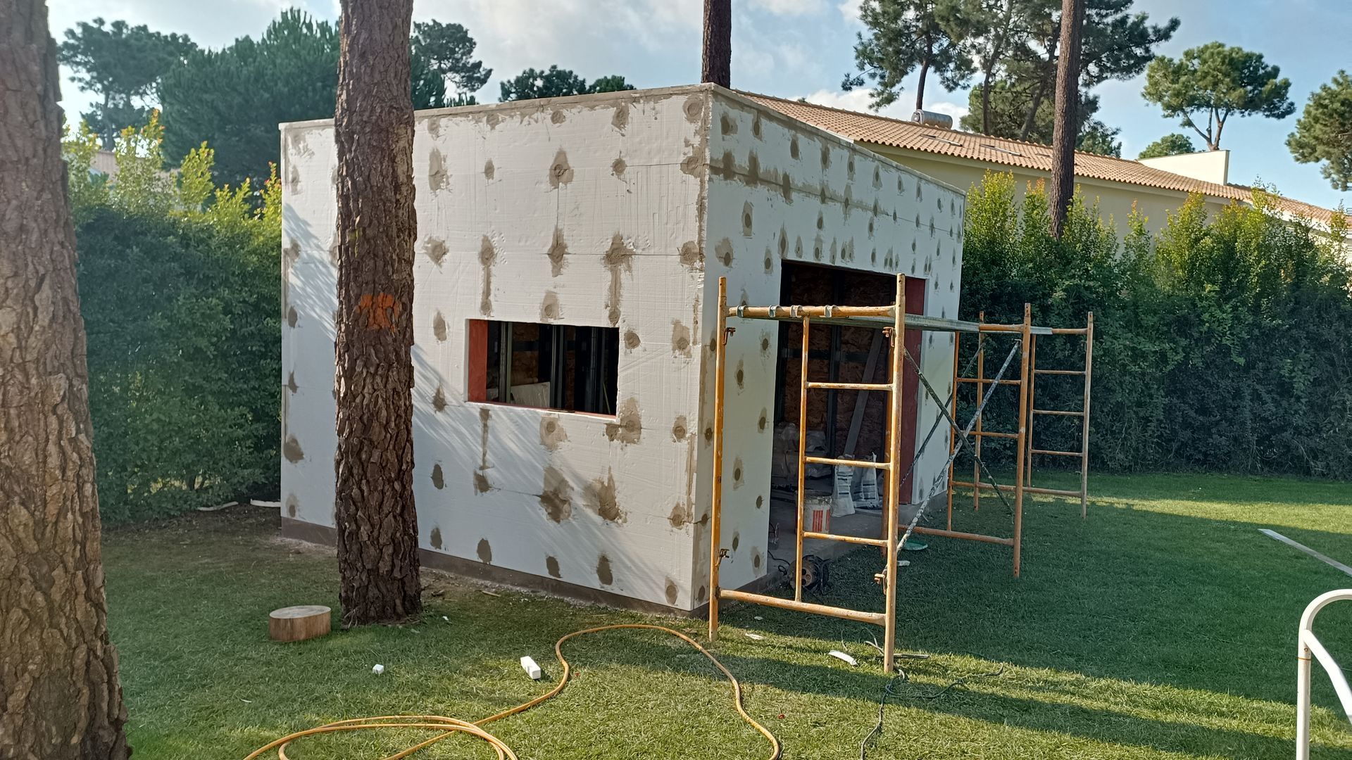 Exterior de um pequeno edifício em construção, com paredes brancas, abertura de janela, andaimes e árvores.