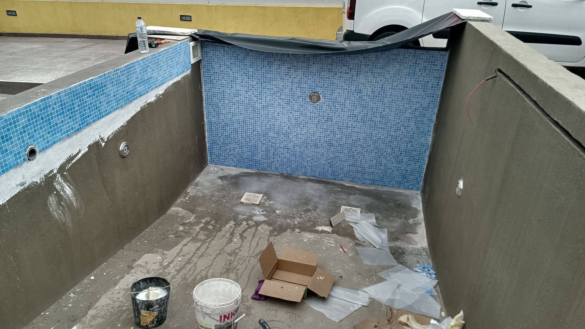 Piscina parcialmente revestida com azulejos em construção; azulejos azuis, concreto cinza, área de trabalho.