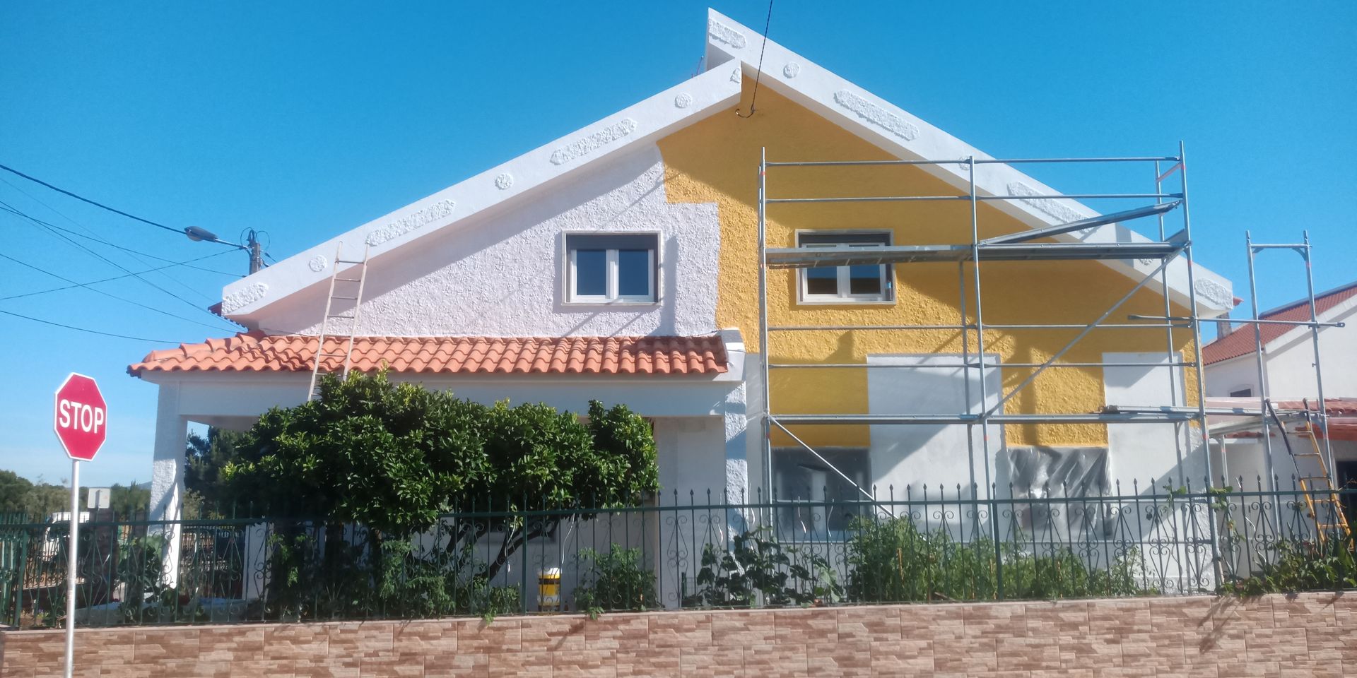 Casa sendo pintada de amarelo; andaimes estão no local. Telhado branco e seção com telhas vermelhas. Placa de pare em primeiro plano.