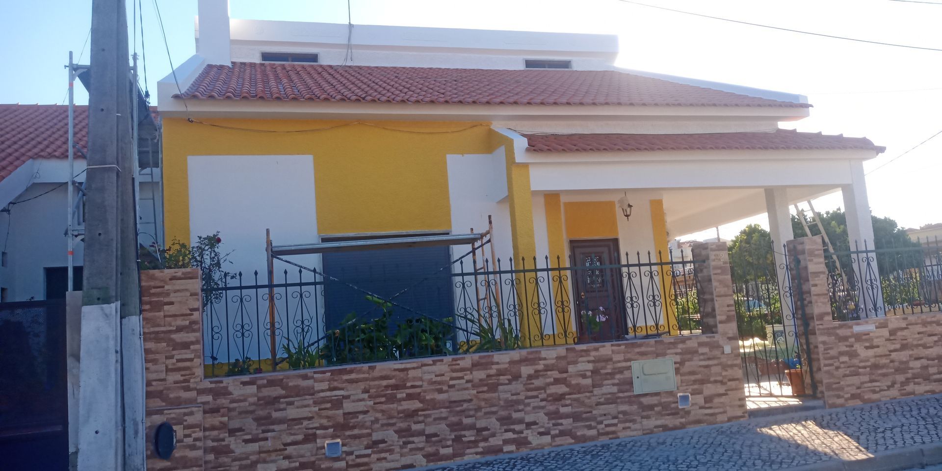 Casa amarela com telhado de telhas vermelhas, parede de tijolos e portão de metal.