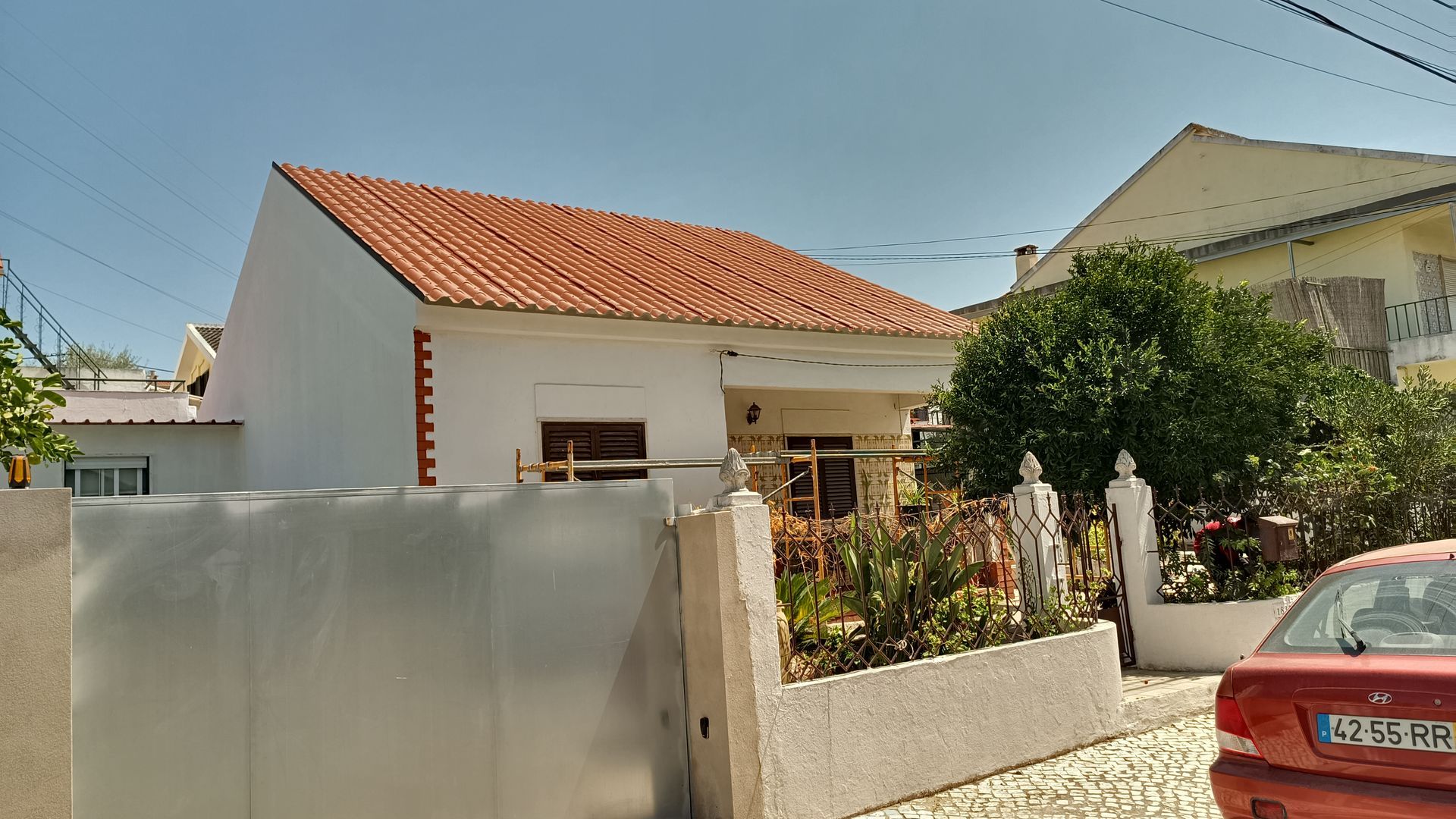 Casa branca com telhado de telha laranja, jardim e um carro vermelho estacionado nas proximidades.