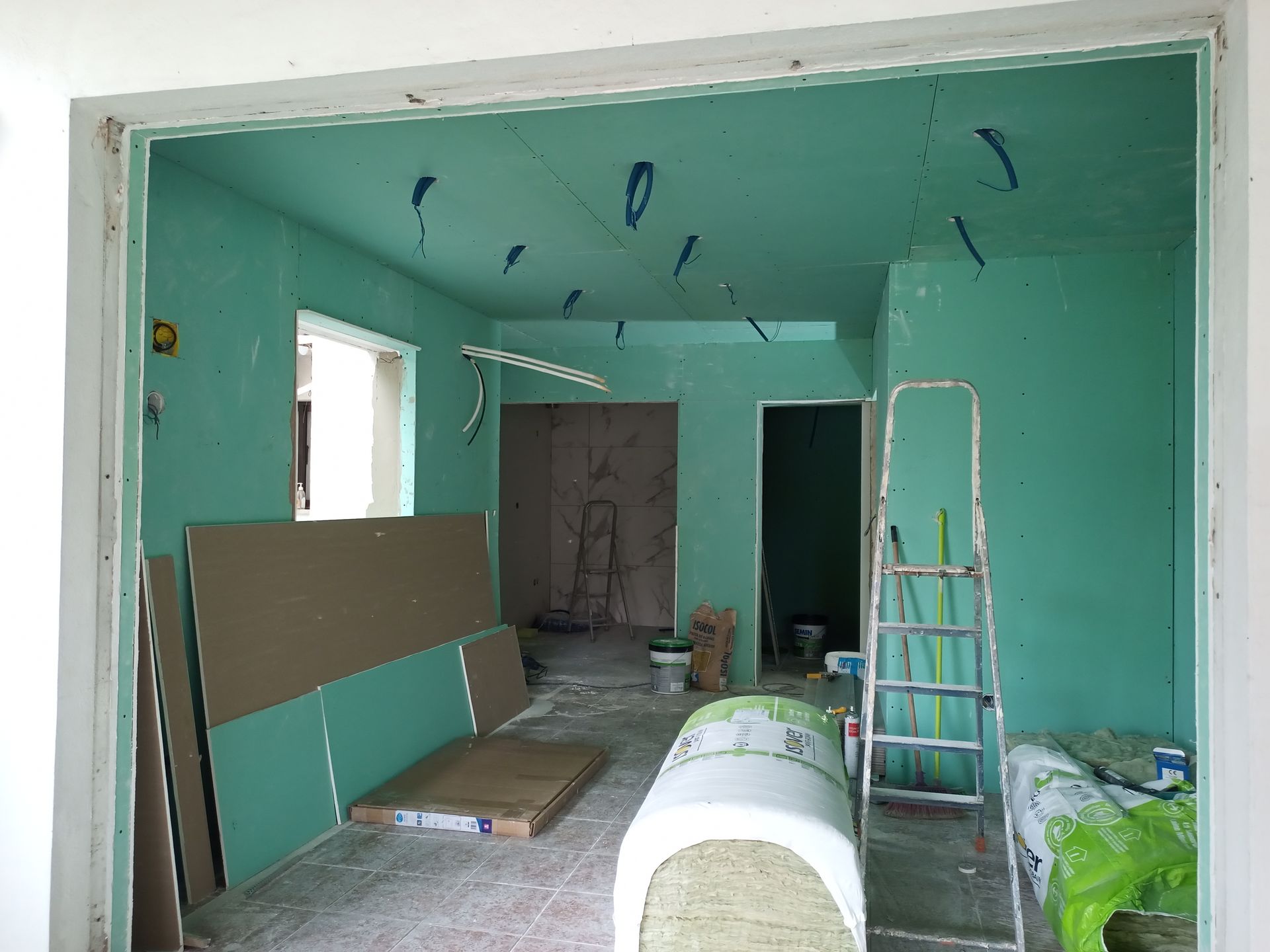 Vista interna de um cômodo em construção com paredes verdes, fiação exposta, drywall e uma escada.