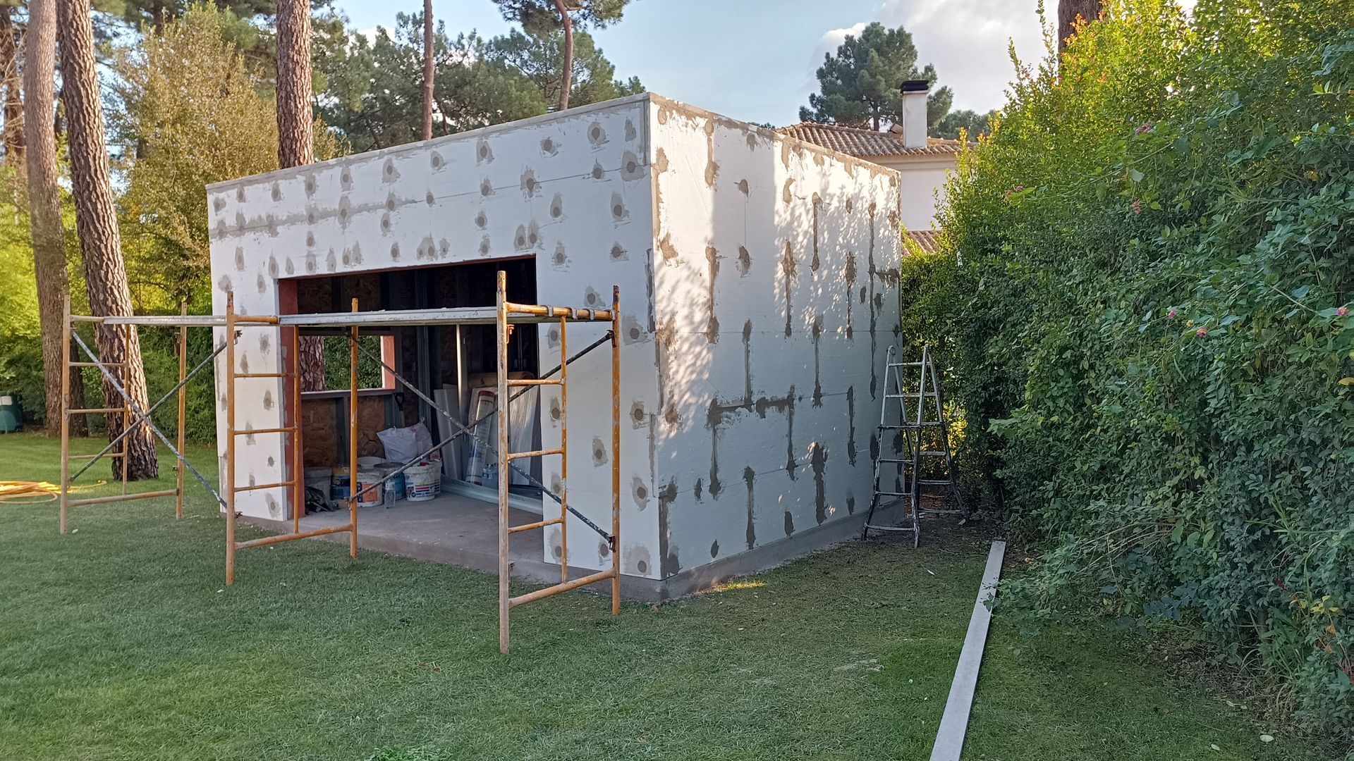 Garagem em construção; paredes externas revestidas com painéis isolantes, andaimes, gramado verde.