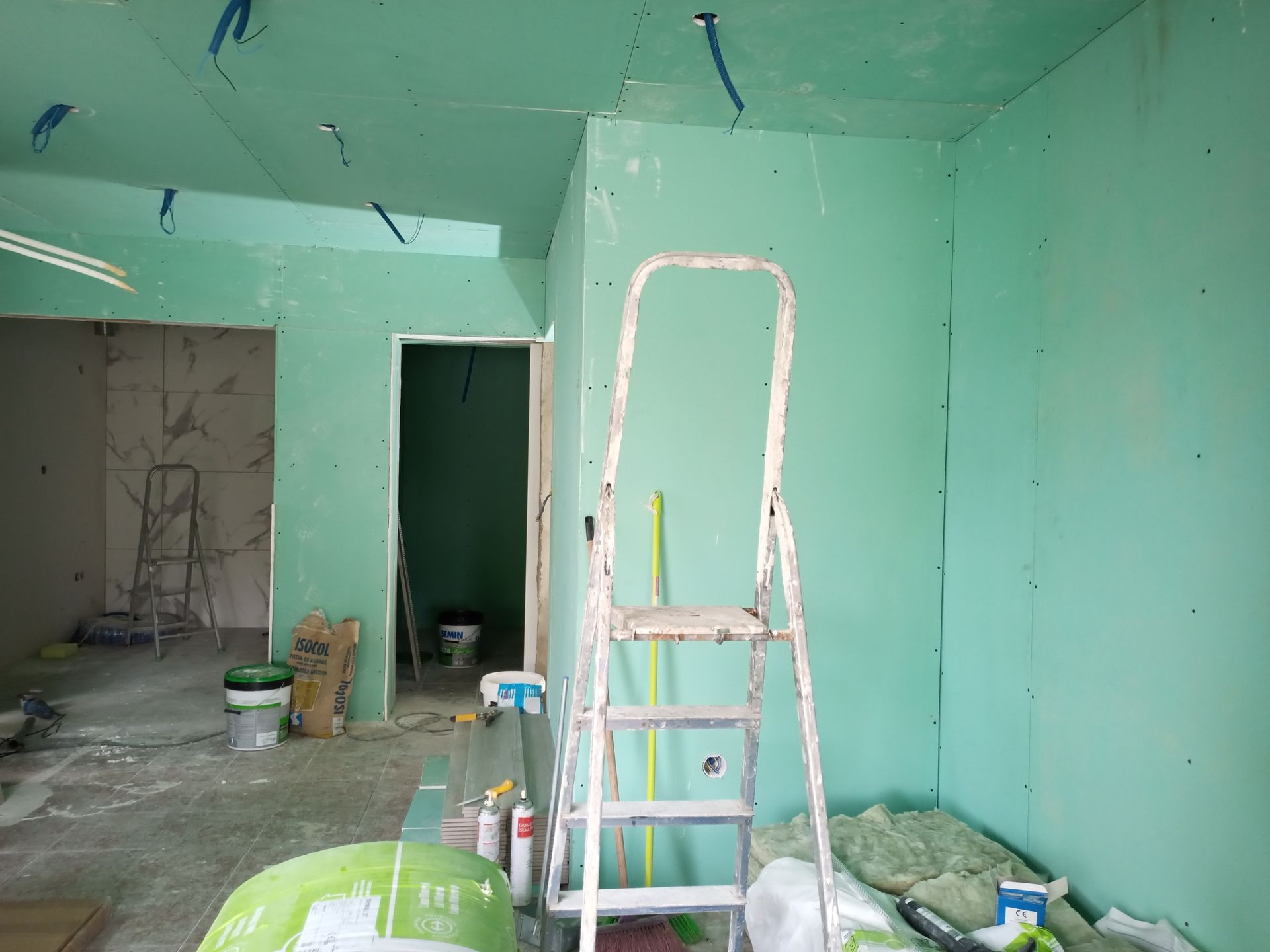 Construção interna: drywall verde, escada, fios expostos, teto inacabado, porta, baldes de tinta, isolamento.