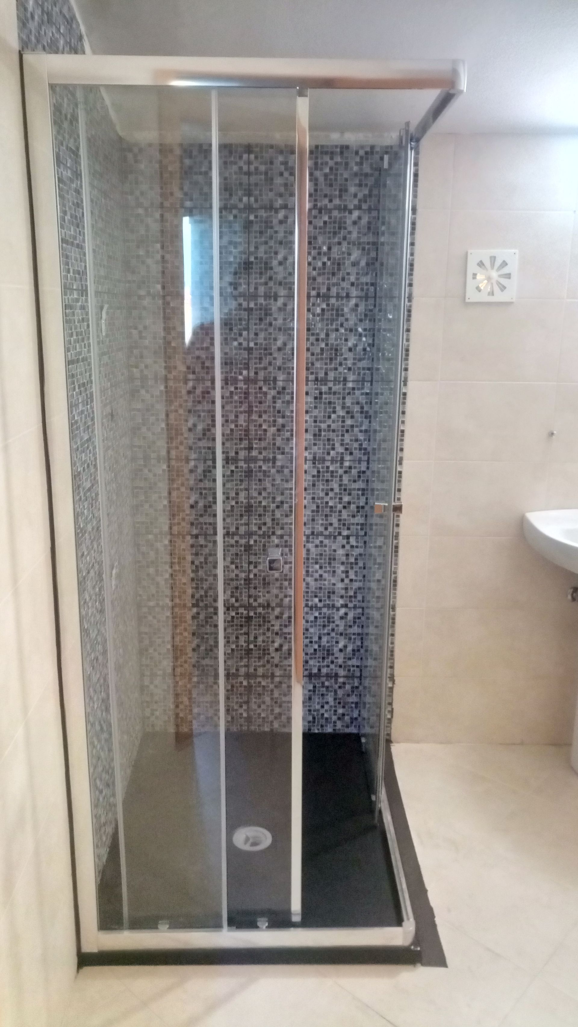 Cabine de duche com portas de vidro, moldura prateada e base escura, encostada a uma parede revestida a azulejos.