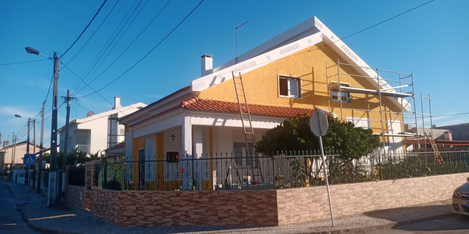 Casa amarela com detalhes brancos e andaimes; telhado de telhas e rua em primeiro plano.