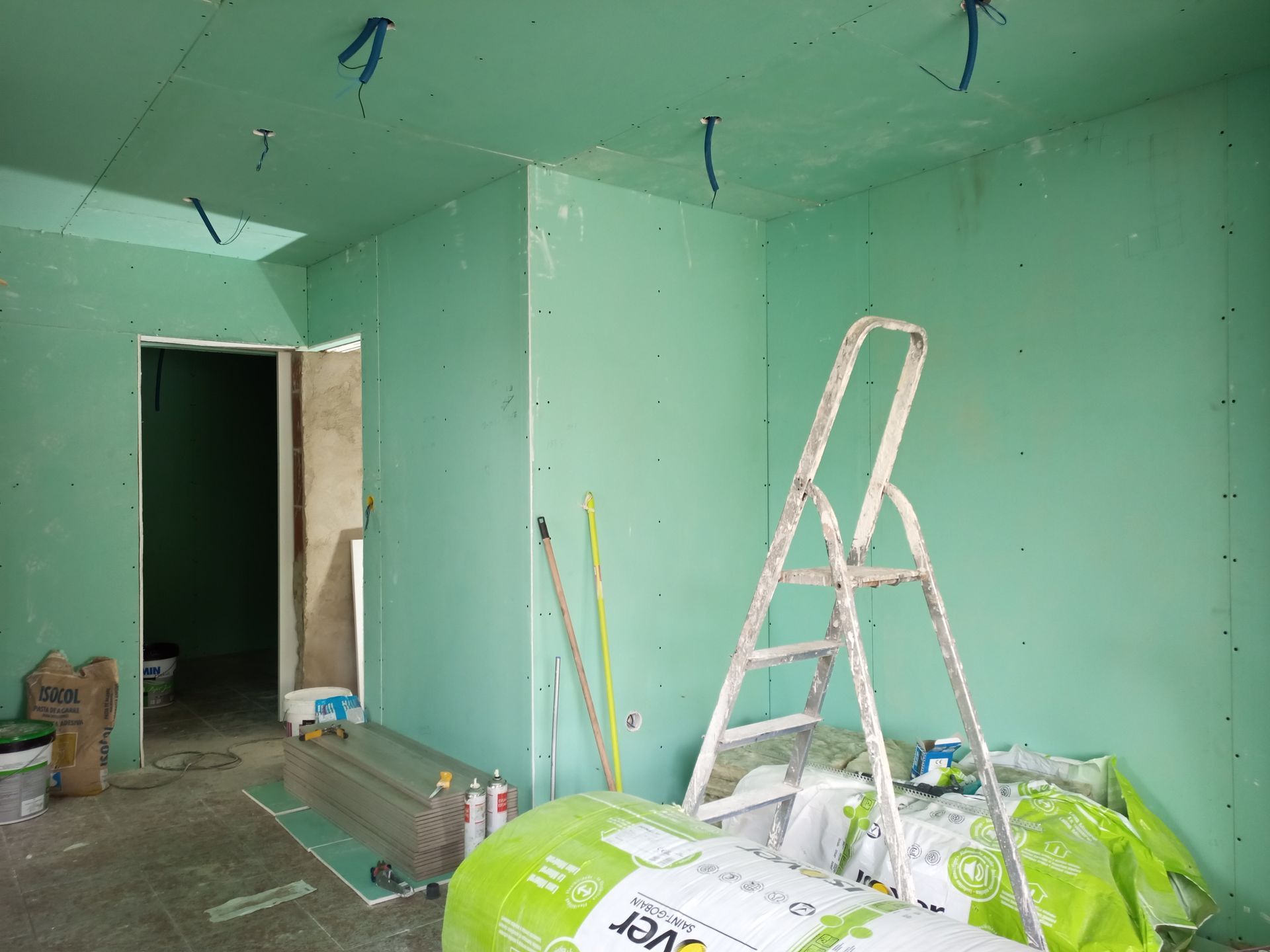 Interior de um cômodo em construção, com drywall verde, escada e materiais de construção.