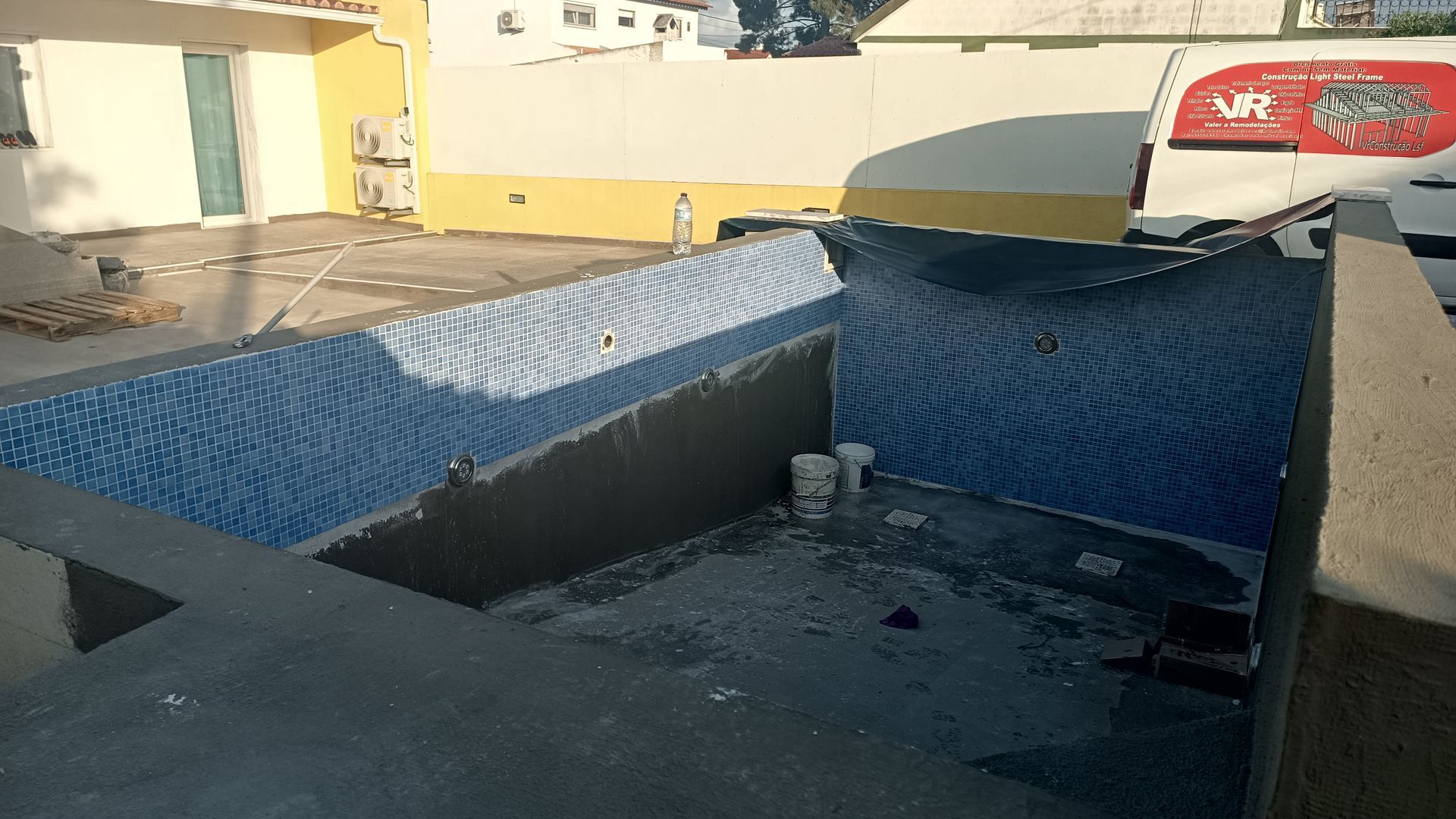 Piscina em construção com paredes revestidas de azulejos azuis, concreto e edifícios circundantes.