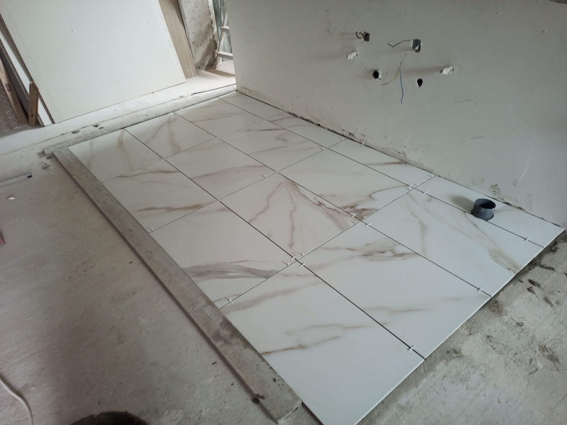 Instalação de azulejos no piso do banheiro em andamento: azulejos brancos com veios marrons, espaçadores e acessórios de encanamento na parede.