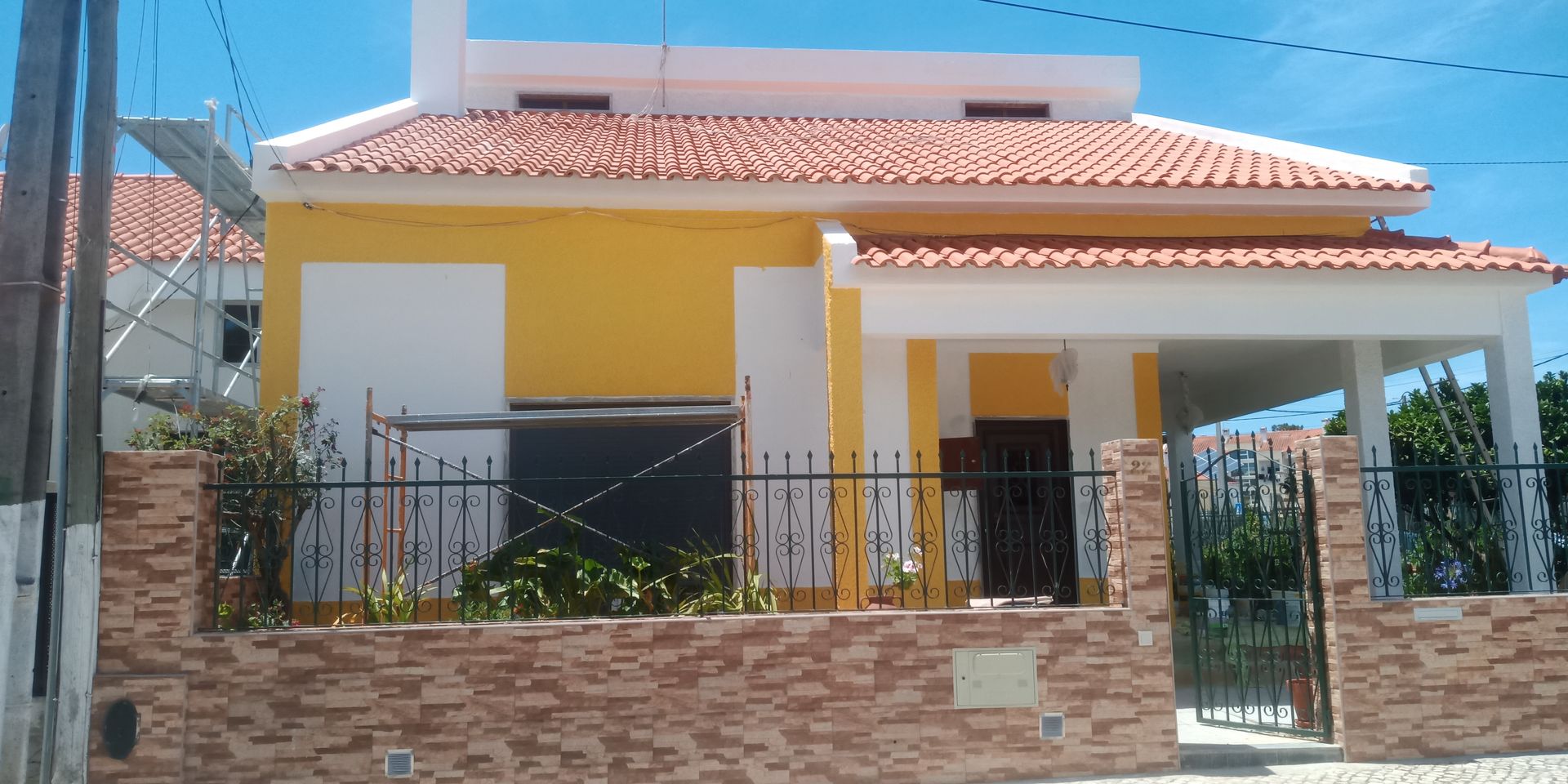 Casa amarela e branca com telhado de telha vermelha, muro de tijolos e portão preto sob um céu azul claro.