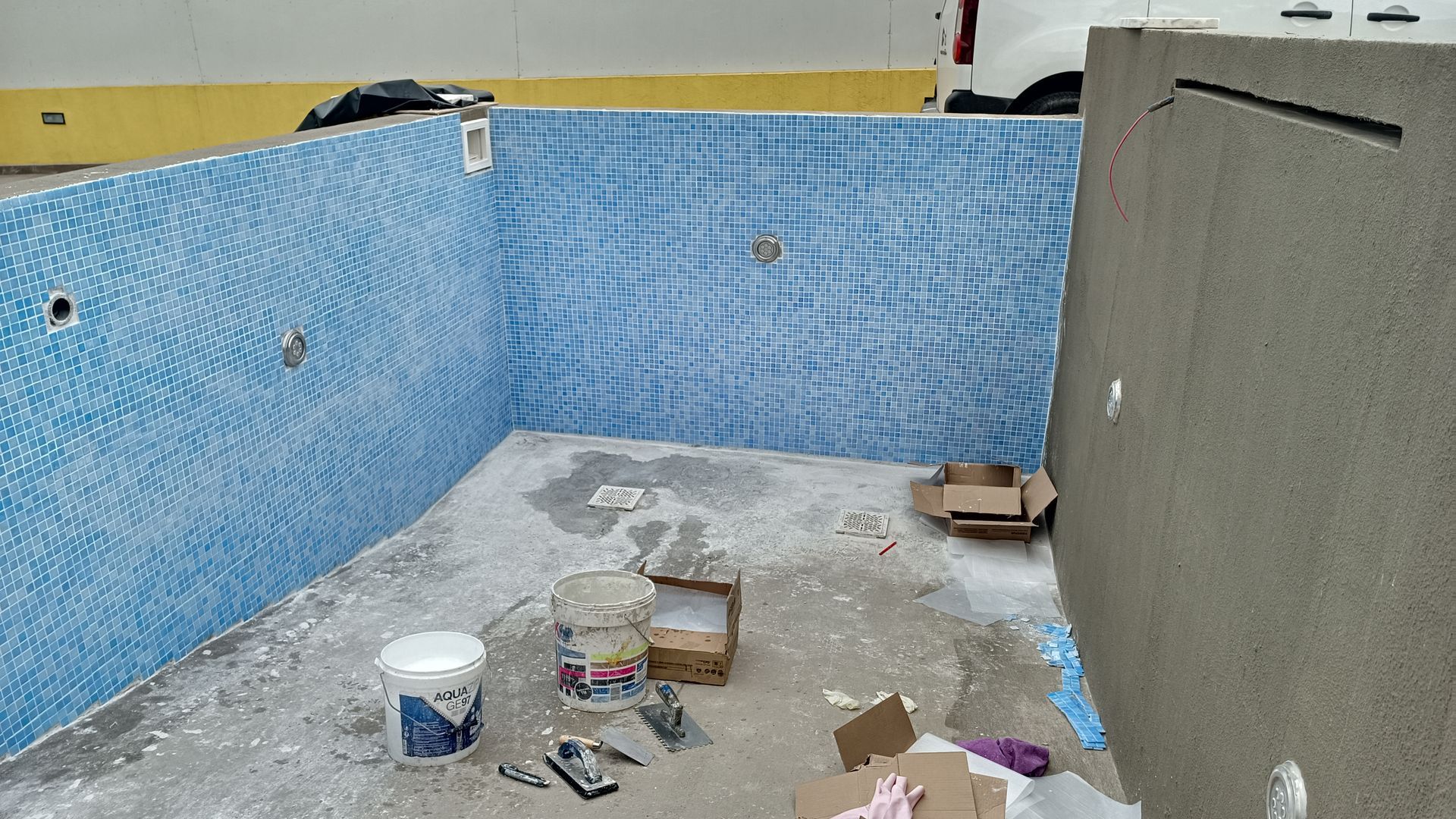 Piscina em construção com paredes de azulejos azuis, baldes brancos e caixas de papelão.