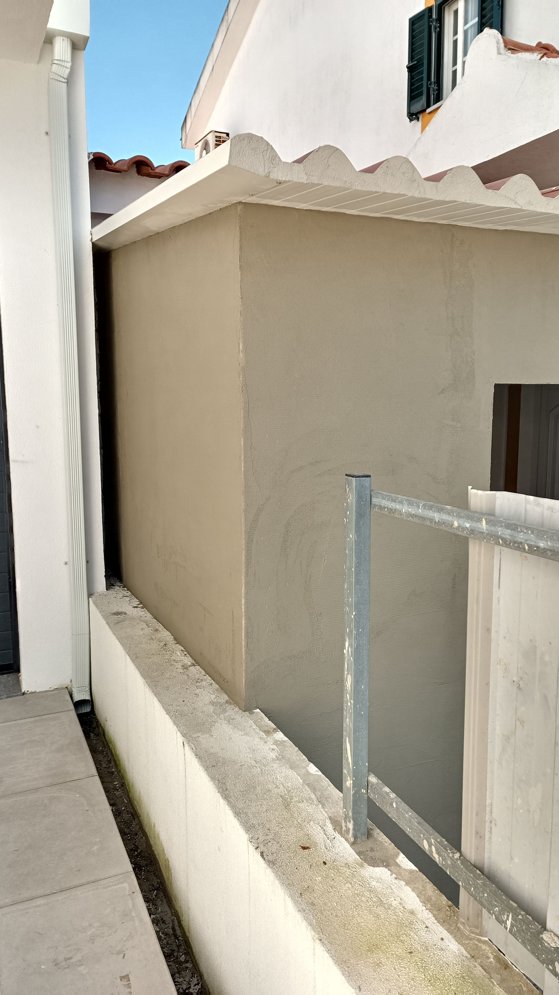 Edifício de estuque bege entre estruturas brancas, com uma grade de metal em primeiro plano.