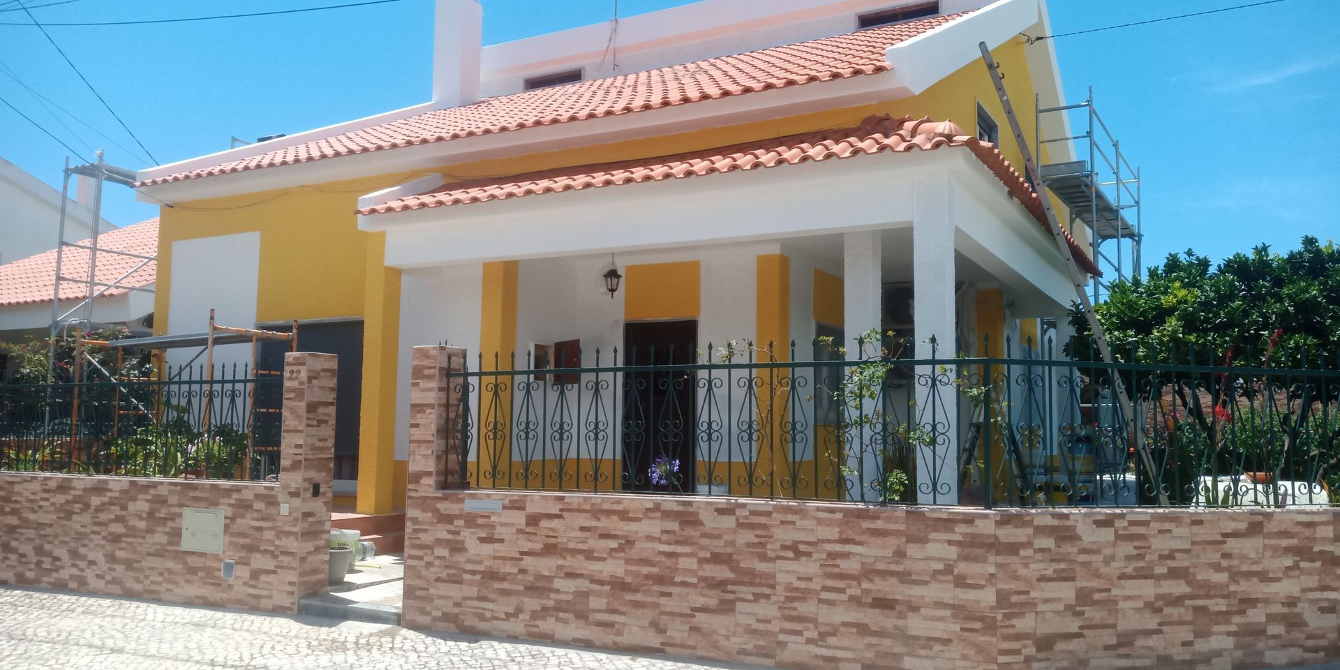 Casa amarela com detalhes brancos, telhado de telha vermelha e muro de tijolos. Andaimes são visíveis na lateral.