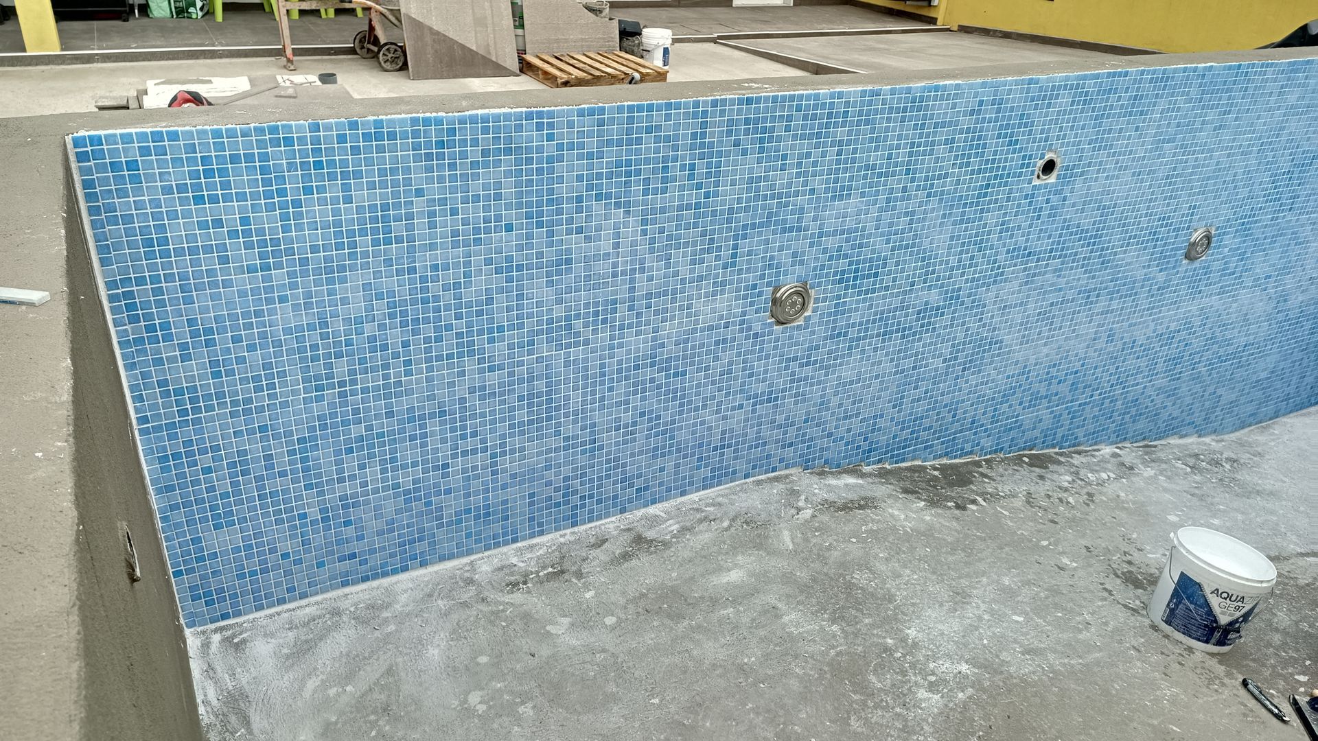 Parede da piscina em construção, com azulejos de mosaico azul instalados e cerca de concreto ao redor.