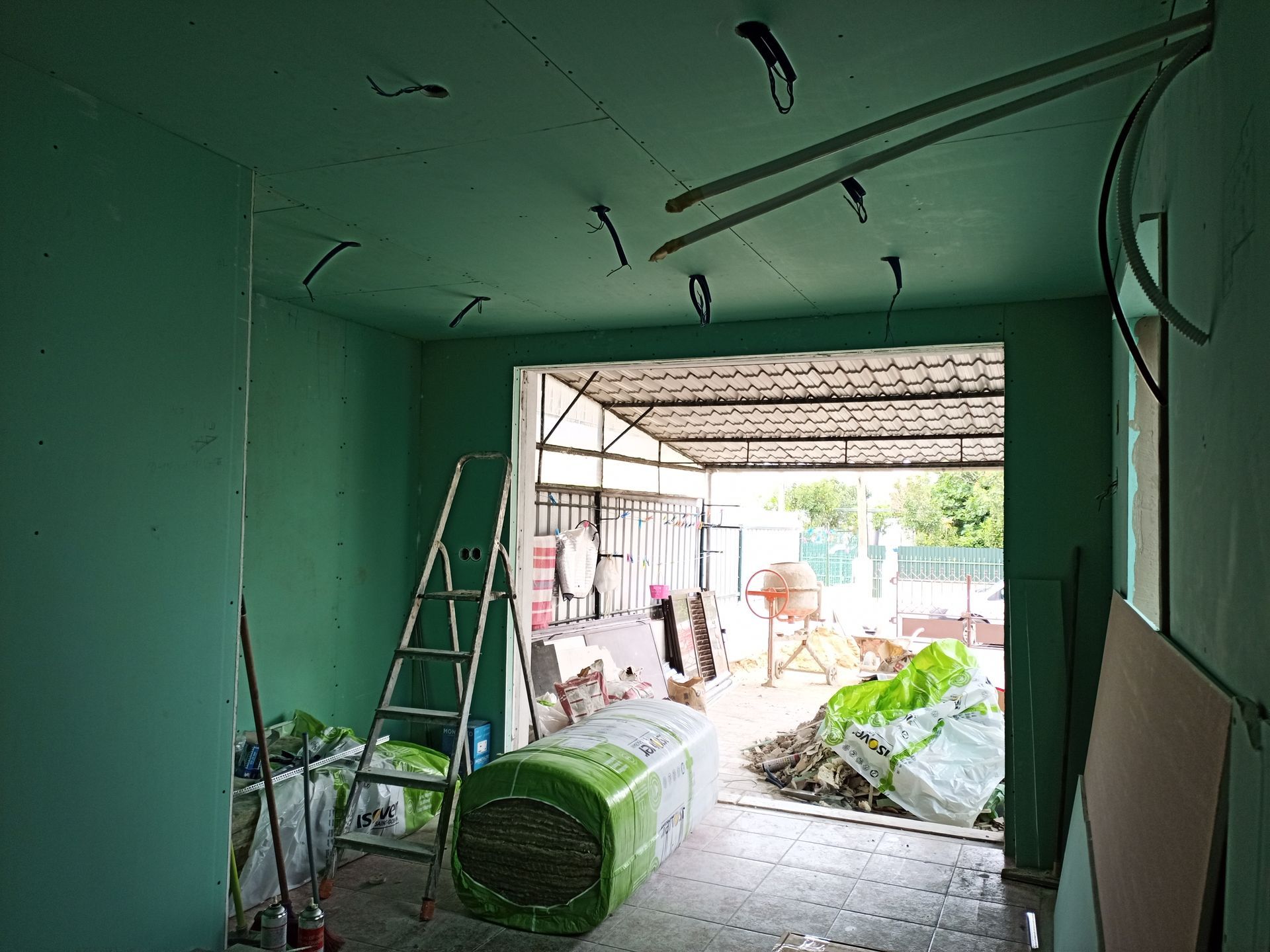 Vista interior de uma sala em construção, com paredes verdes, uma escada e uma porta aberta para uma área externa.