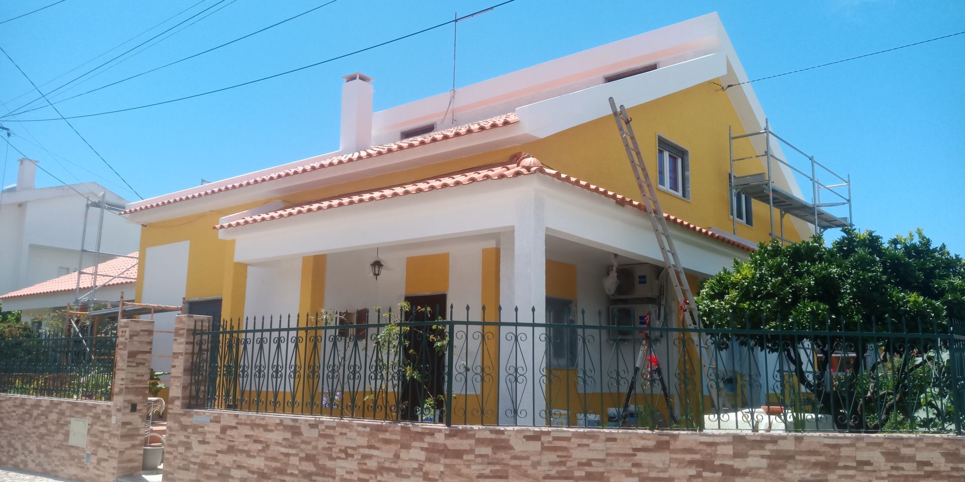 Casa amarela com detalhes brancos, telhas vermelhas, cerca preta, parede de tijolos e céu azul.