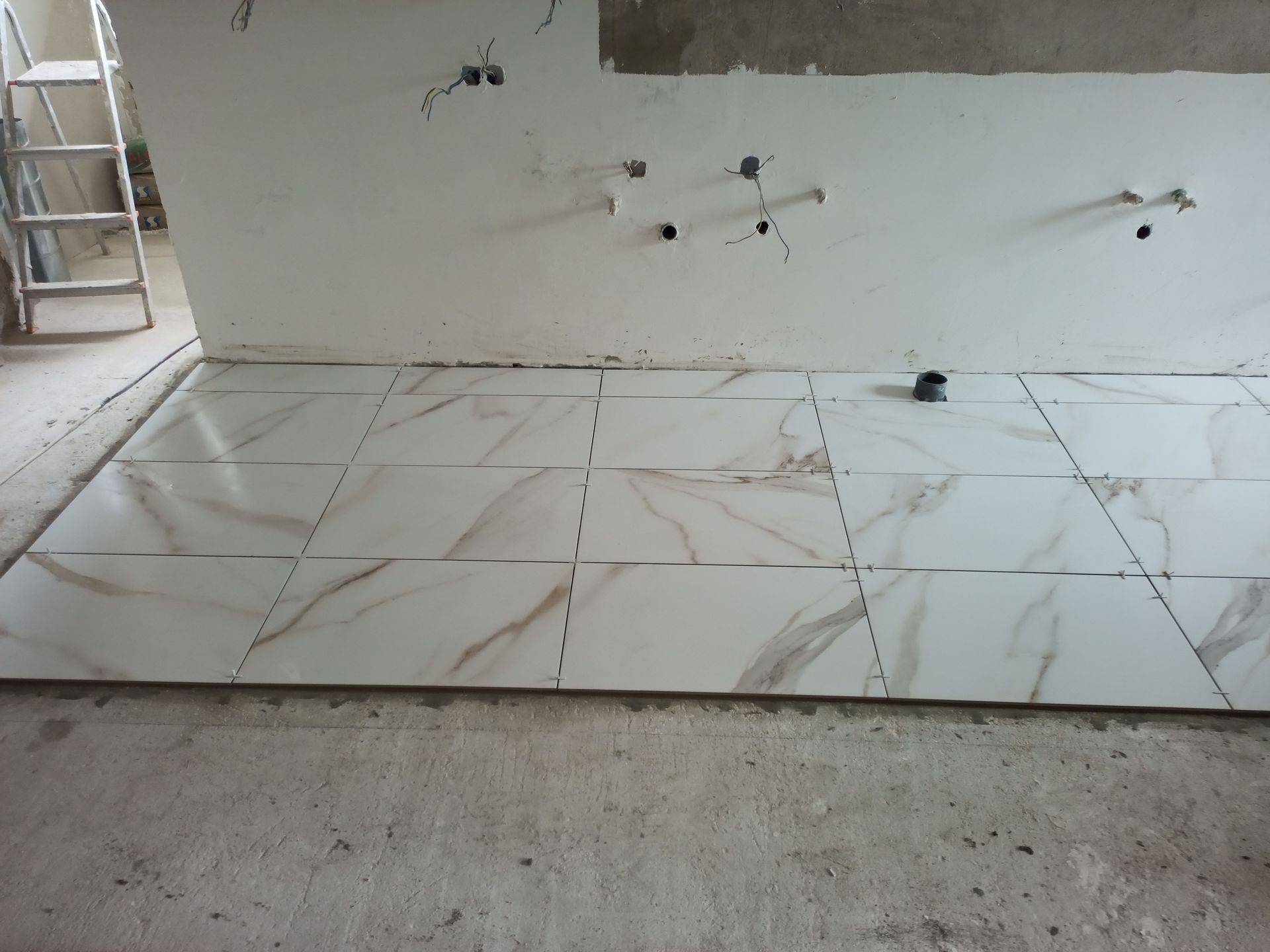 Azulejos brancos com aparência de mármore assentados sobre um piso de concreto próximo a uma parede com tomadas elétricas e uma escada ao fundo.