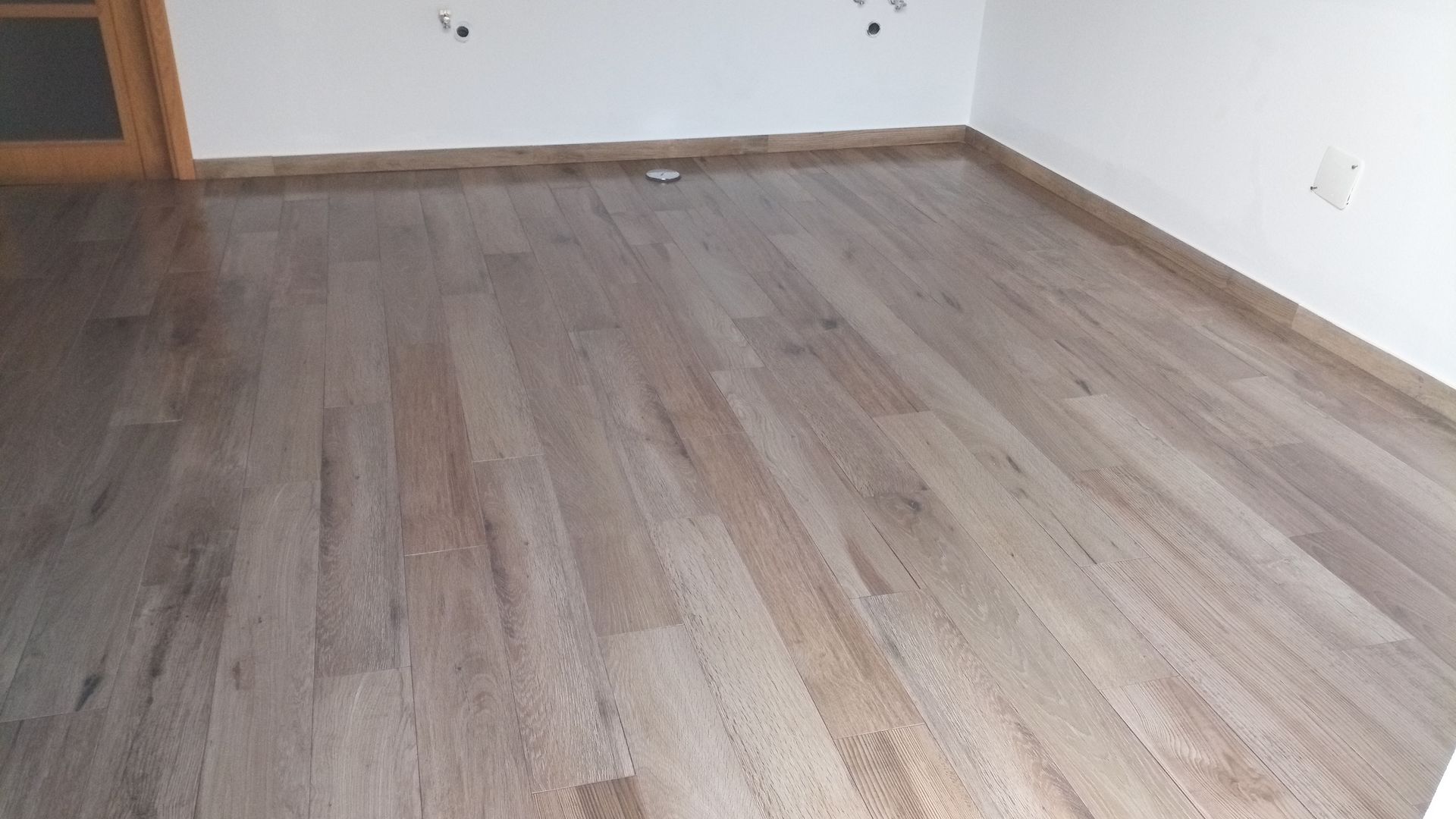 Piso de madeira em um cômodo com paredes brancas, detalhes em marrom e uma porta visível à esquerda.