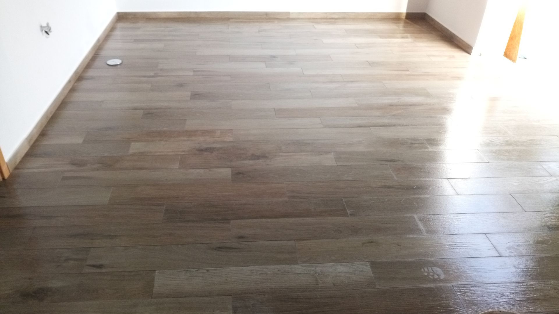 Piso de cerâmica imitando madeira em um cômodo com paredes brancas. A luz do sol entra por uma porta à direita.