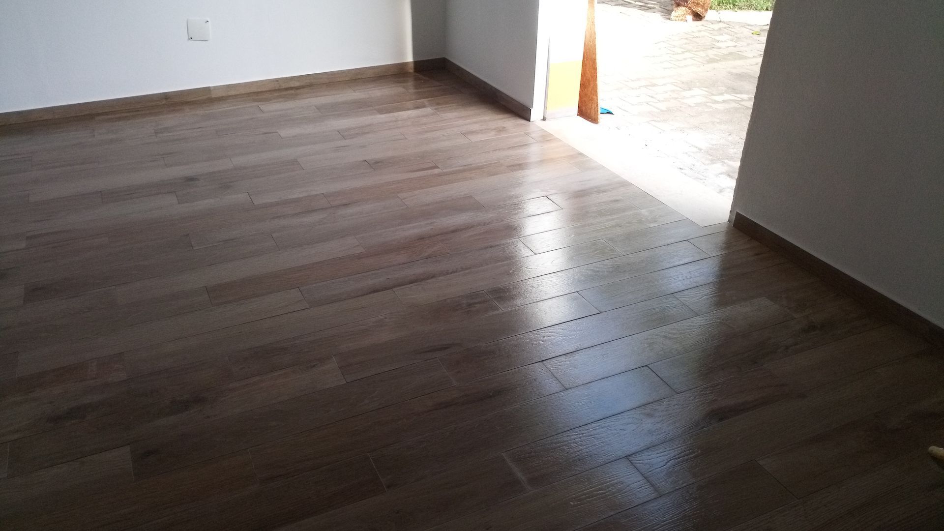 Piso de madeira em um cômodo com luz solar entrando por uma porta aberta. As paredes são brancas com detalhes em tom escuro.