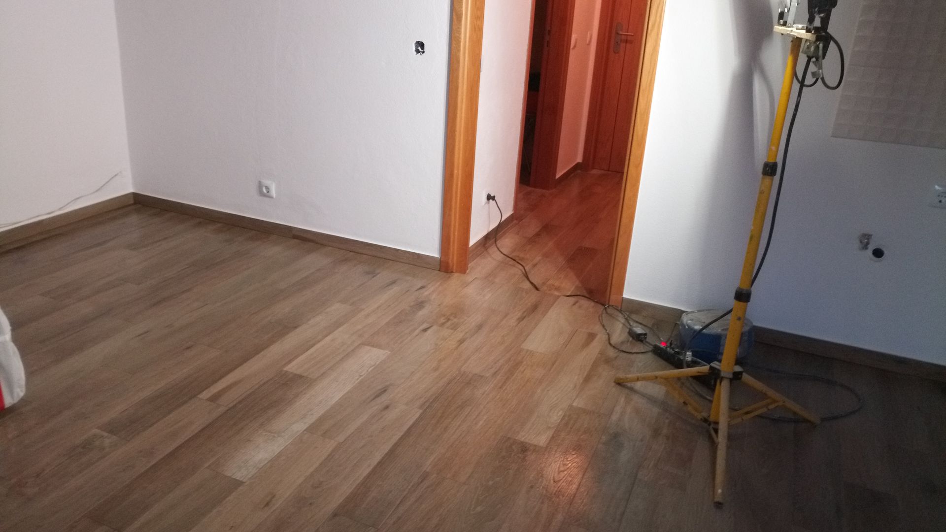 Sala com piso que imita madeira, paredes brancas e porta aberta. Há uma luminária de trabalho instalada no local.