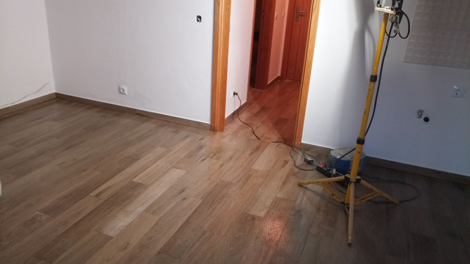 Sala com piso de madeira, paredes brancas, porta e luminária de trabalho.