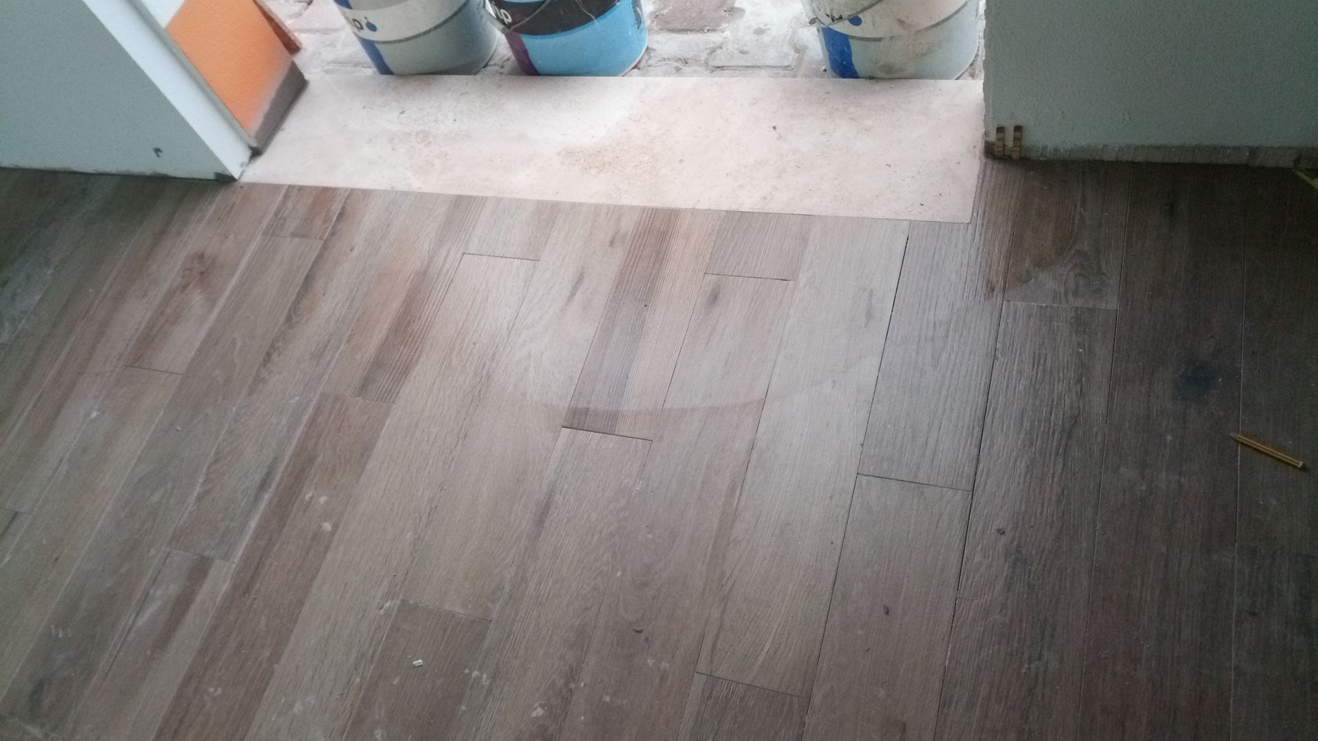 Piso de madeira maciça, parcialmente instalado, próximo a uma porta e baldes. O piso é marrom claro.