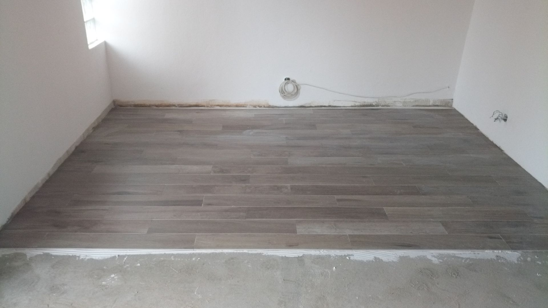 Quarto com piso de porcelanato imitando madeira. Paredes brancas, uma pequena janela e tomada elétrica visíveis. Rodapé de concreto na borda do piso.