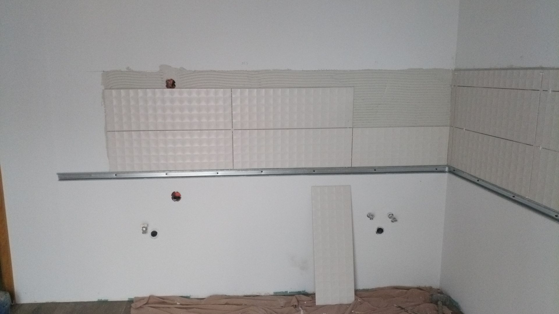 Parede da cozinha em construção, parcialmente revestida com azulejos. Azulejos brancos em contraste com paredes brancas.