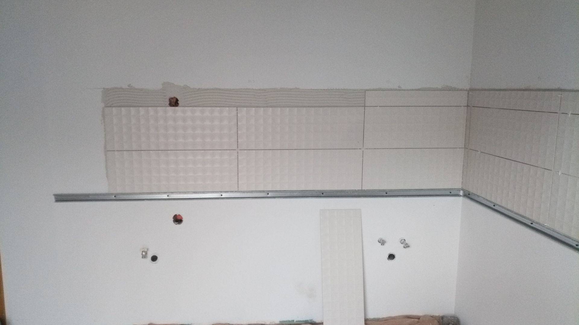 Revestimento de azulejos brancos em andamento, guia metálica instalada e tomadas elétricas expostas na parede branca.