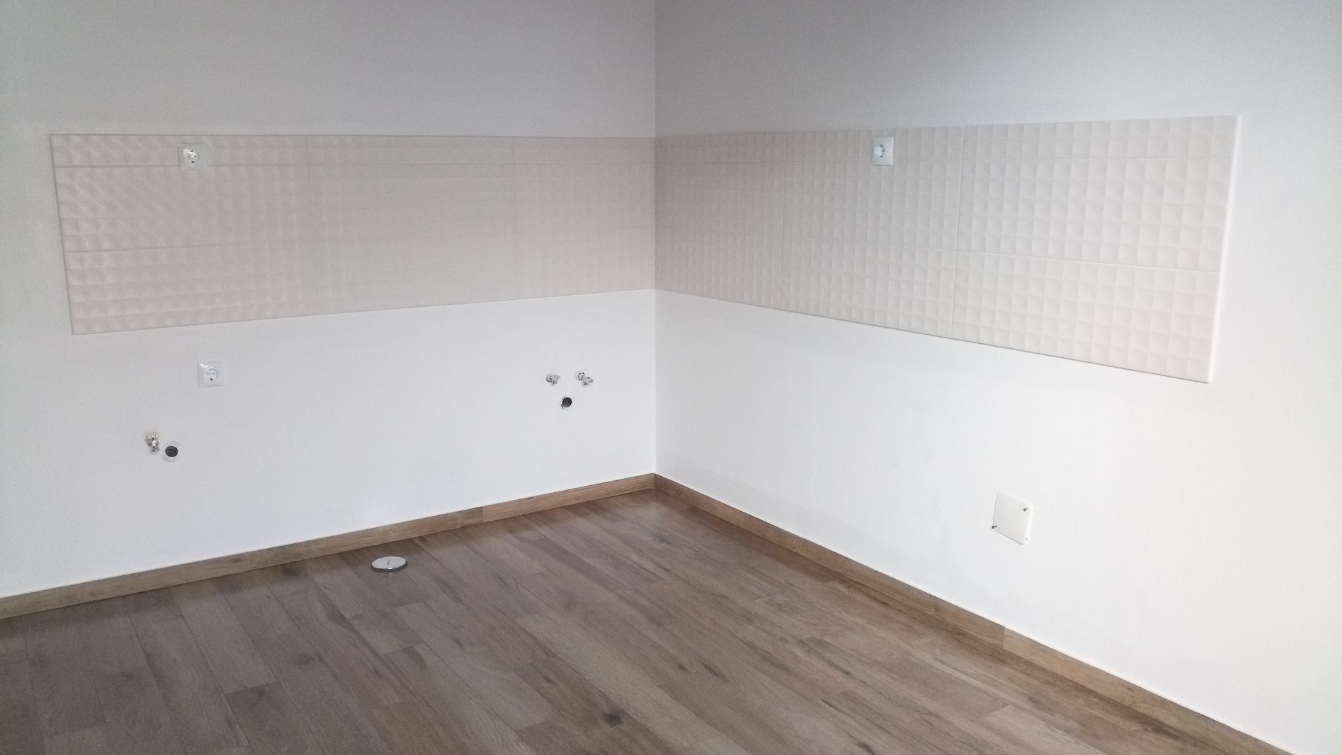 Quarto vazio com piso imitando madeira e paredes brancas. Um canto possui dois painéis bege na parede. Tomadas elétricas são visíveis.