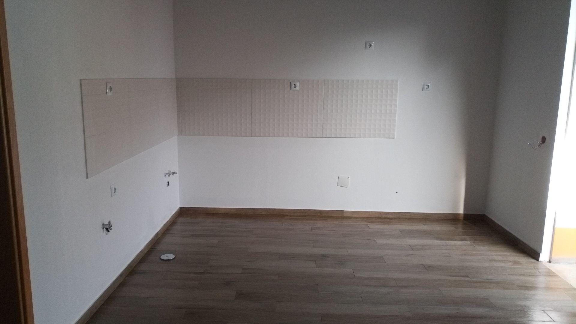 Sala vazia com piso de madeira clara e paredes brancas. Um quadro branco e um painel perfurado estão fixados na parede. A luz do sol entra.