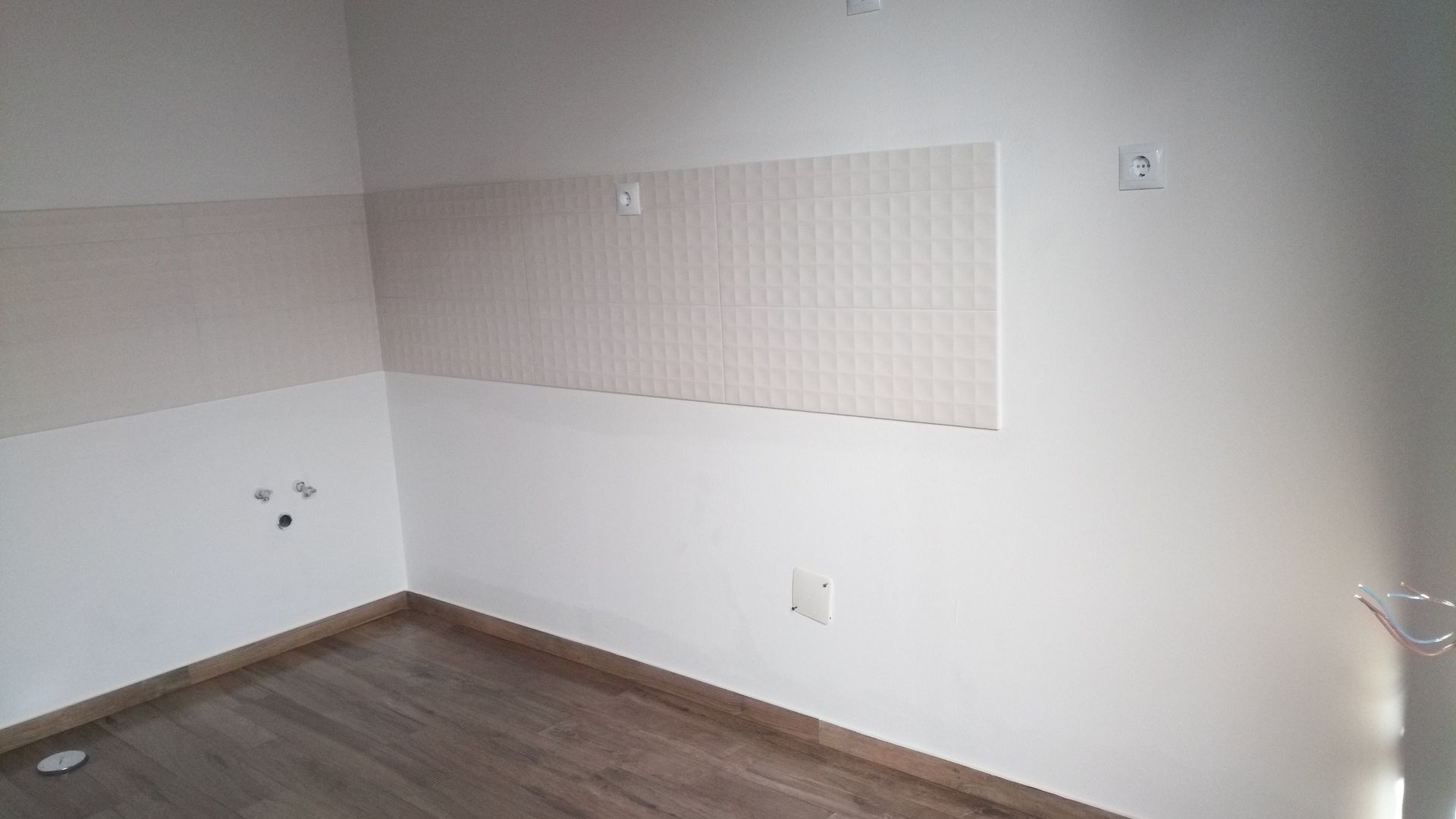 Sala vazia com piso imitando madeira, paredes brancas e painéis decorativos. Tomadas elétricas visíveis.