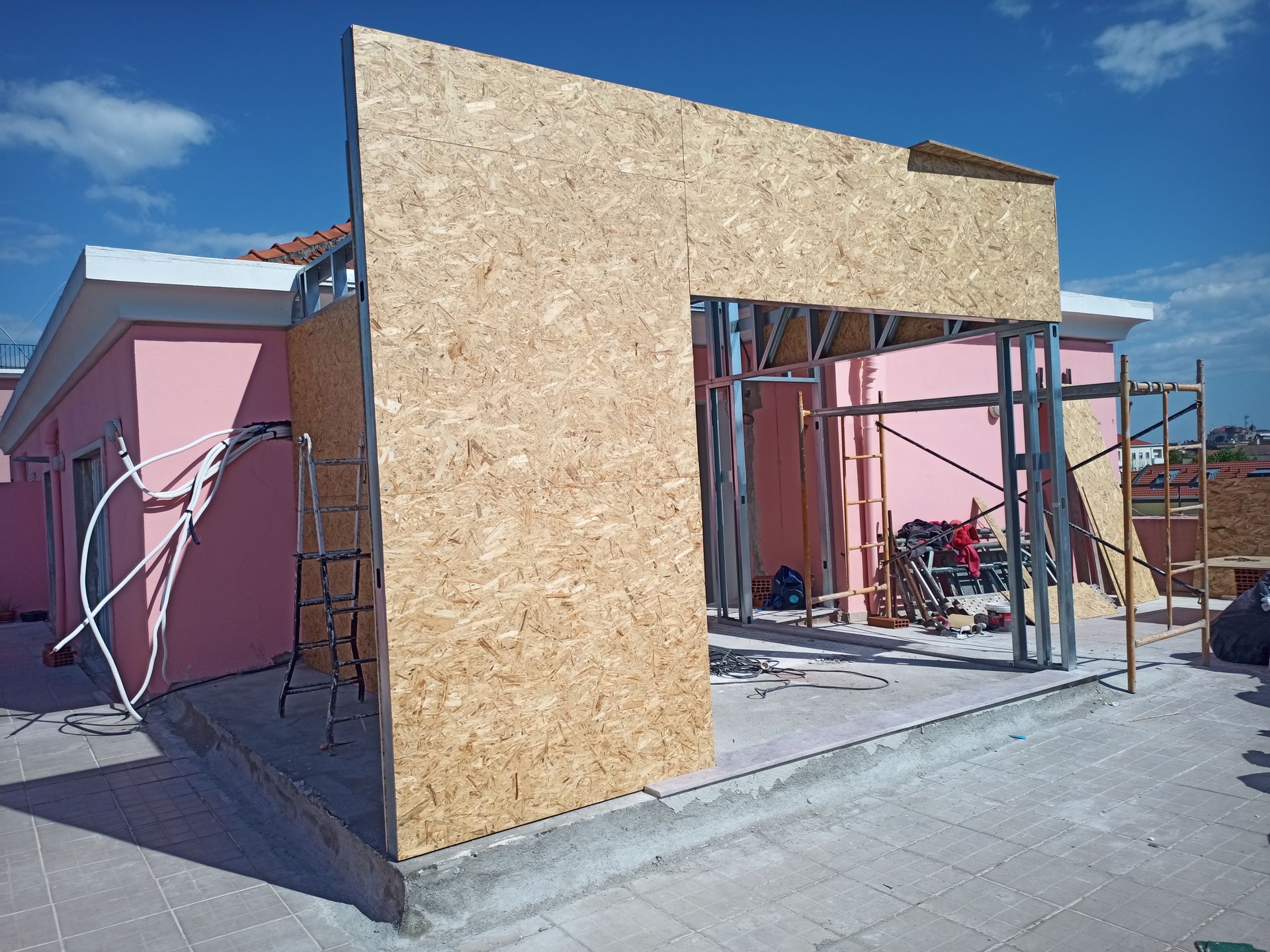 Construção de uma parede com estrutura metálica e painéis de madeira em um telhado. Um prédio rosa aparece ao fundo.