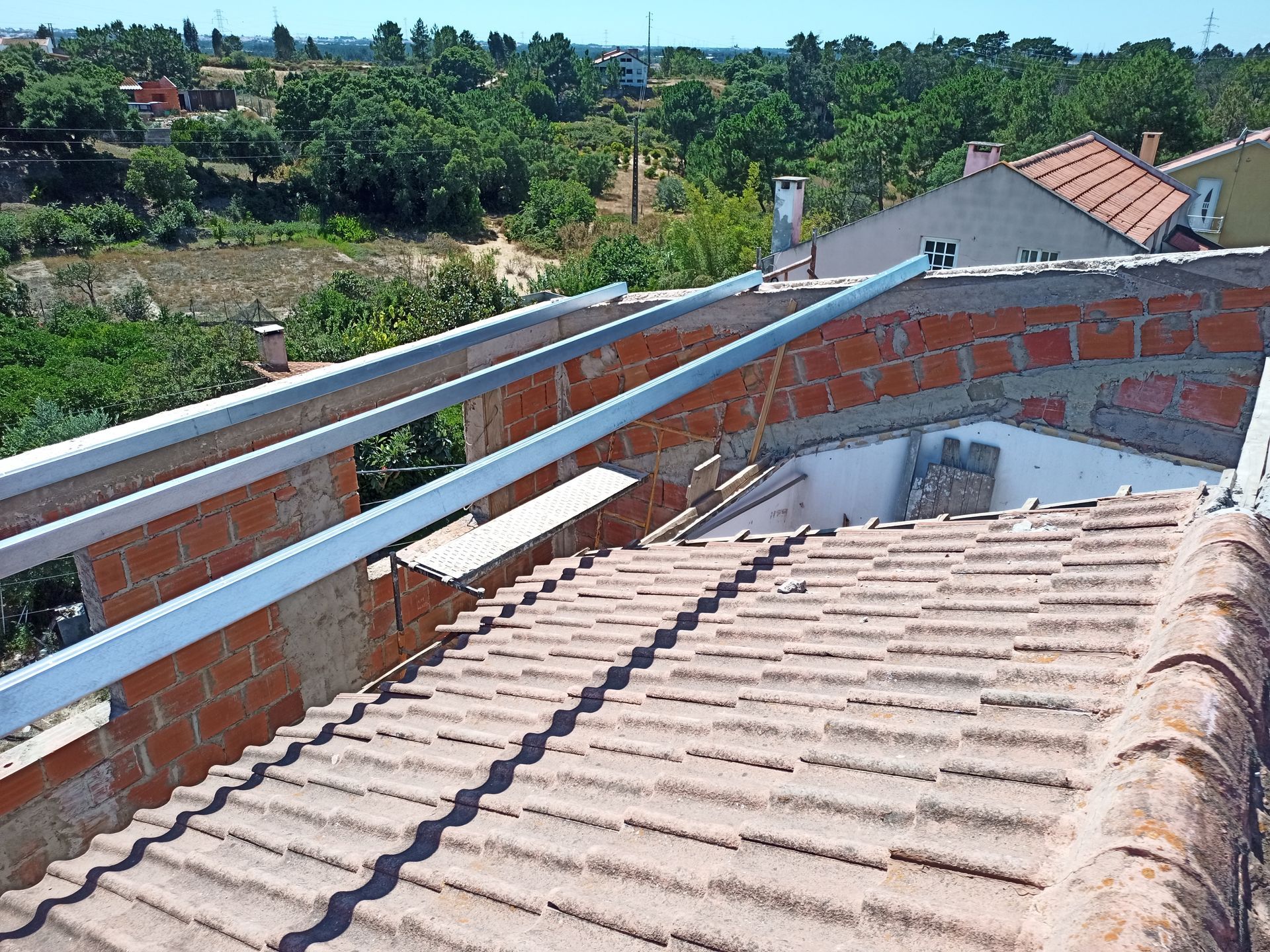 Telhado com telhas vermelhas, alvenaria e estrutura metálica, tendo como pano de fundo árvores e céu azul.