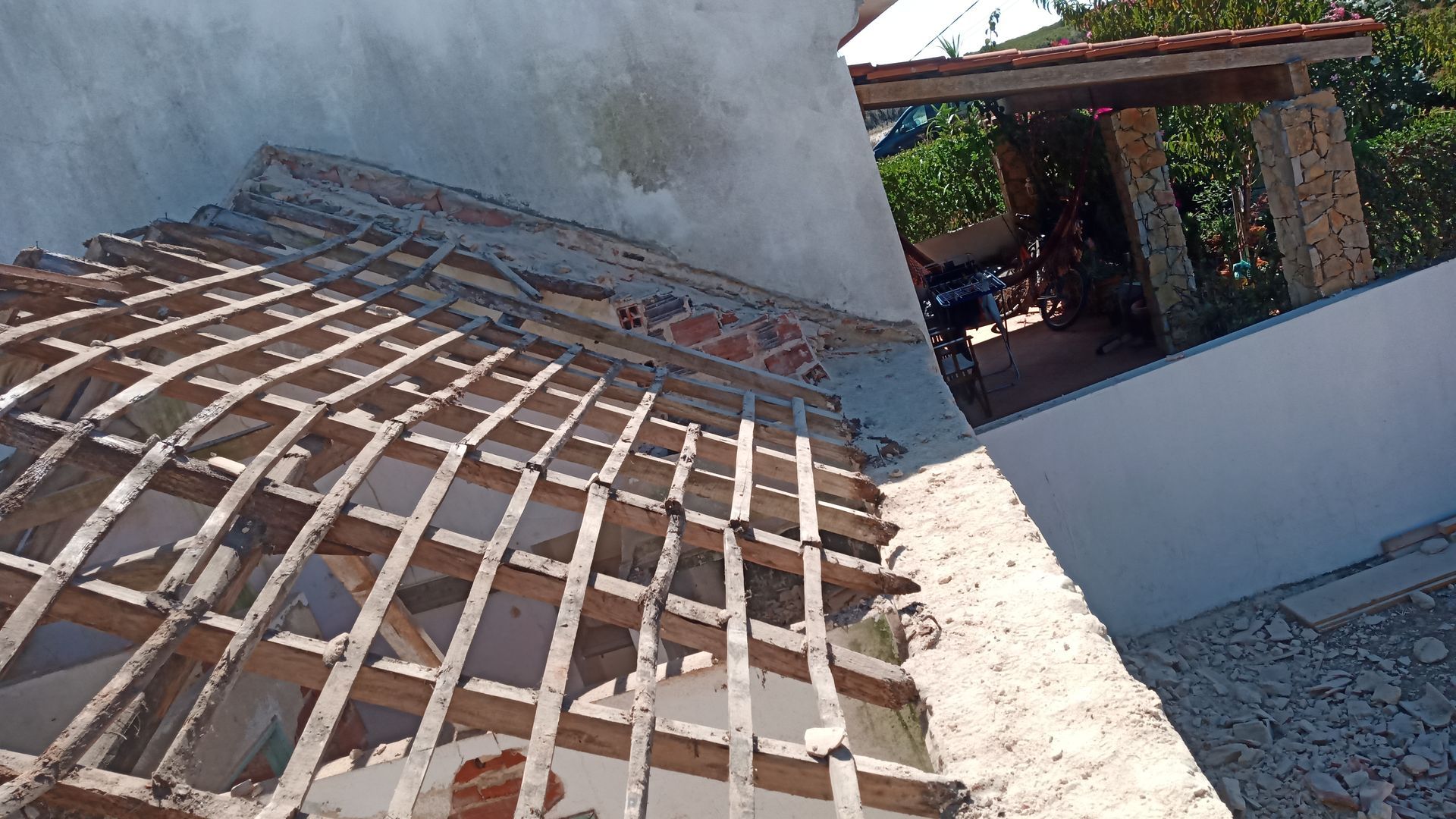 Estrutura do telhado demolida com vigas de madeira expostas, ao lado de uma parede branca e um pátio externo.
