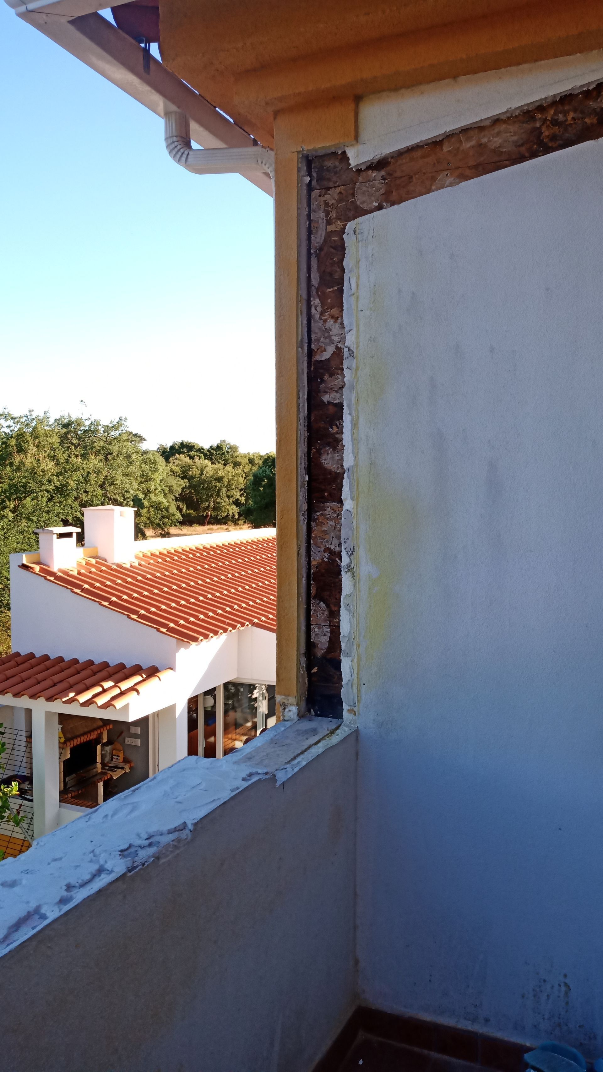 Vista da varanda: Janela aberta revelando tijolos, gesso e uma casa com telhado de terracota, sob um céu claro.