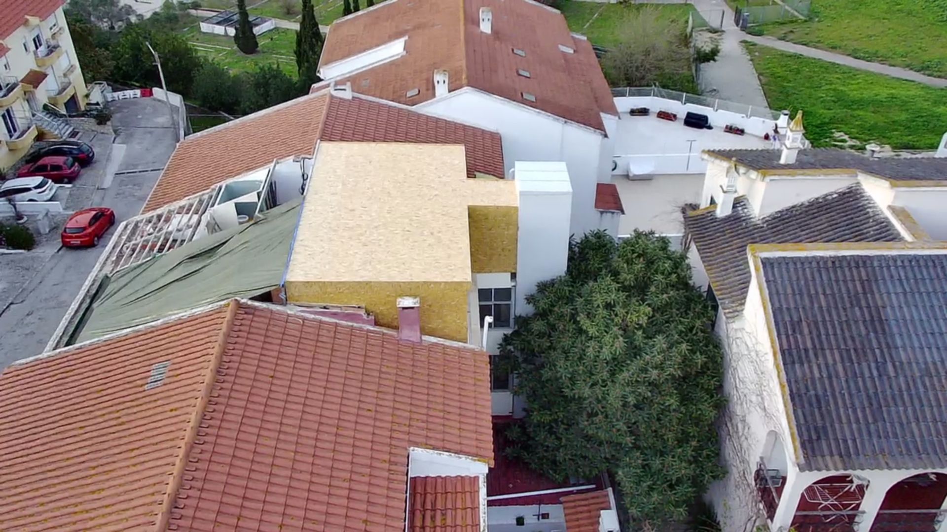 Vista aérea de vários edifícios com telhados vermelhos e bege, uma árvore verde e uma rua com carros estacionados.