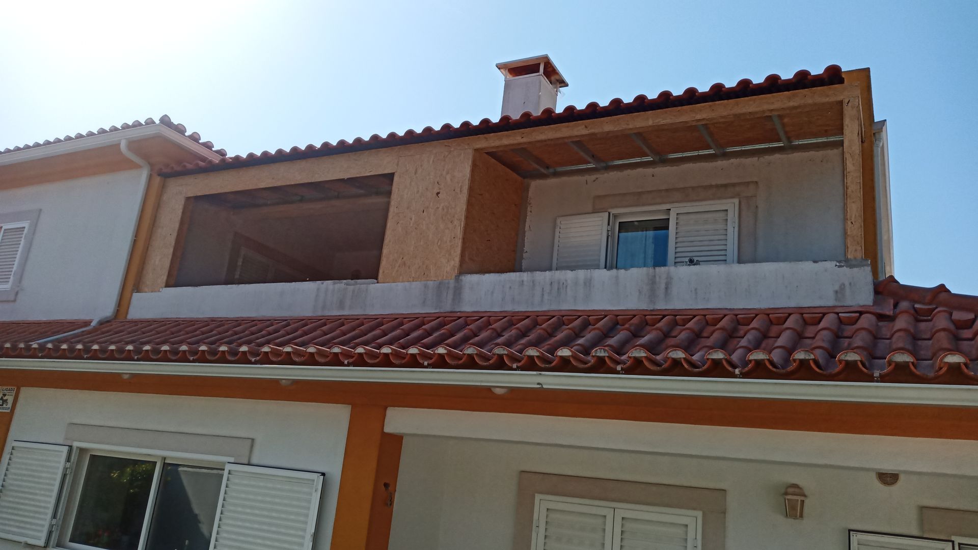 Exterior do edifício com telhado de telha vermelha, varanda em construção e persianas brancas.