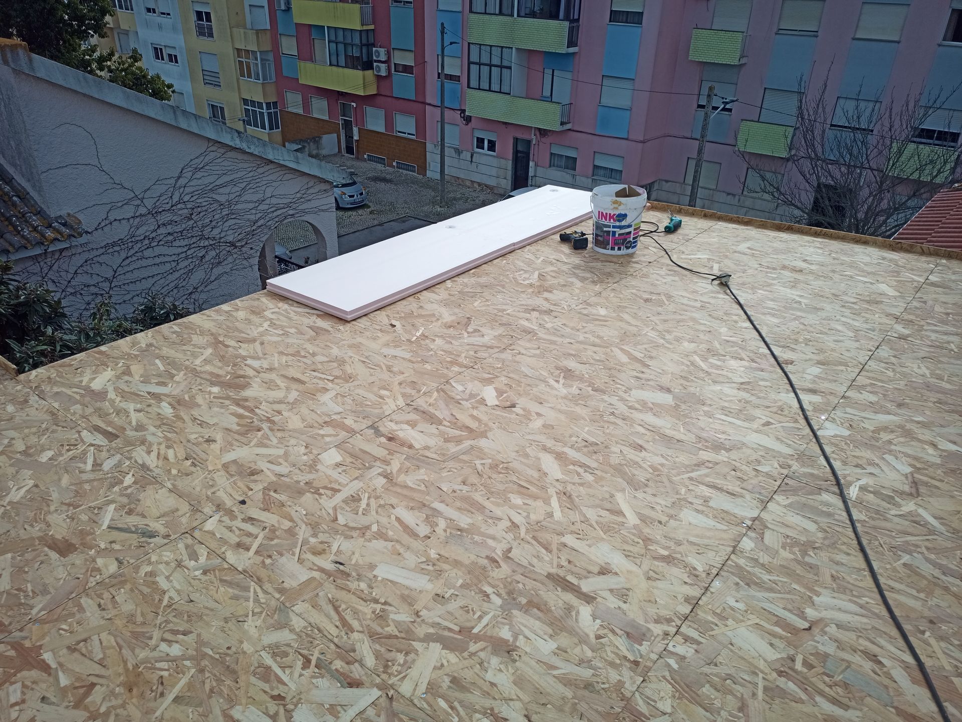 Telhado com painéis de madeira, placa de isolamento, balde de tinta e cabos, tendo a cidade como pano de fundo.