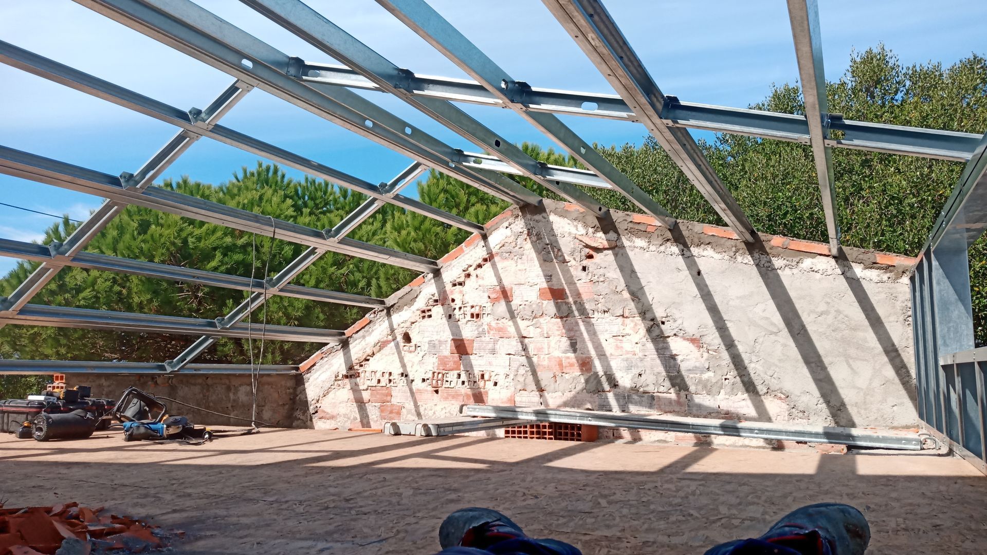 Estrutura metálica do telhado sobre uma parede de tijolos desgastada, com árvores ao fundo sob um céu azul.
