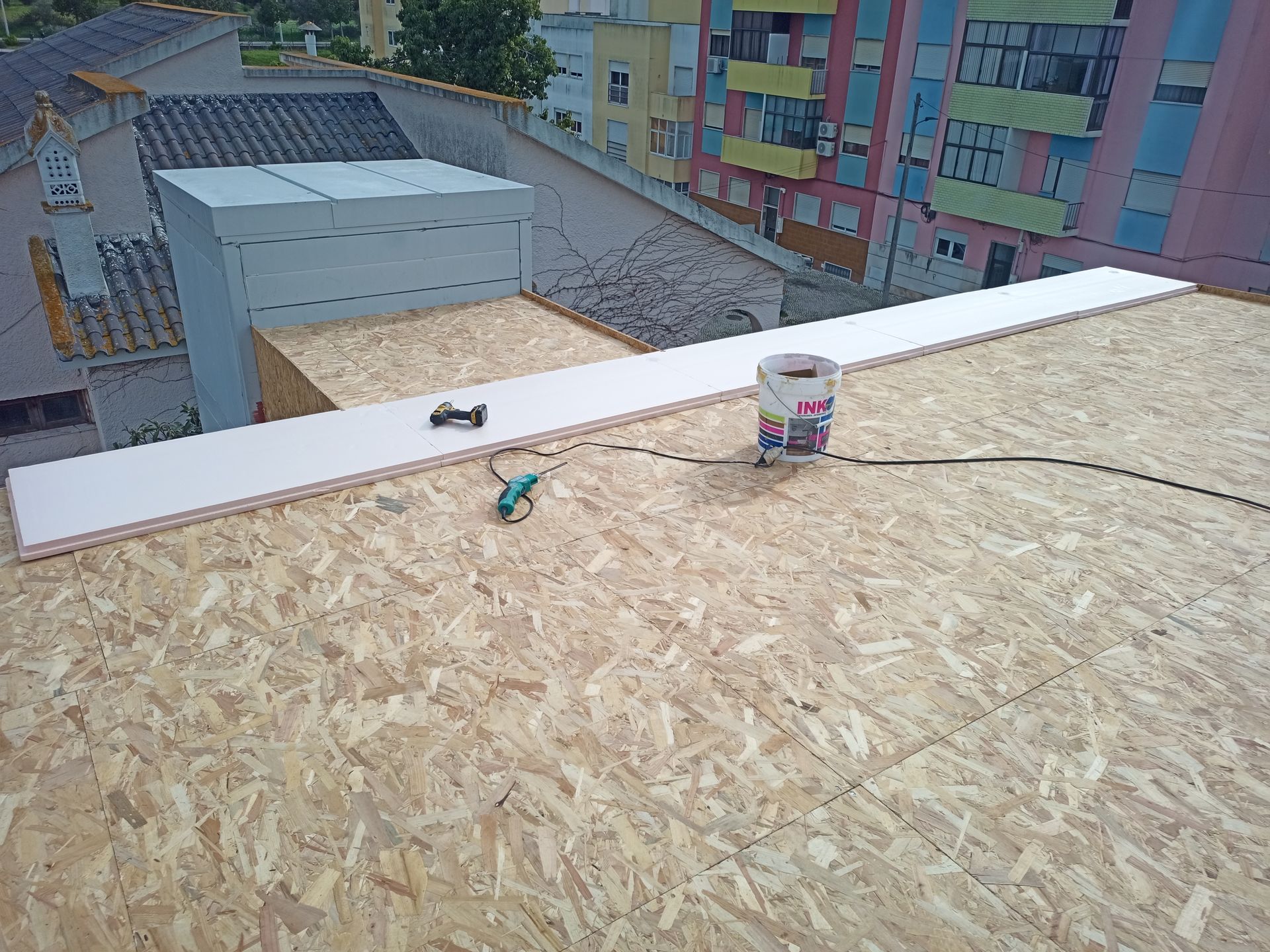 Telhado com materiais de construção: uma longa tábua branca, um balde e ferramentas. Prédios ao fundo.