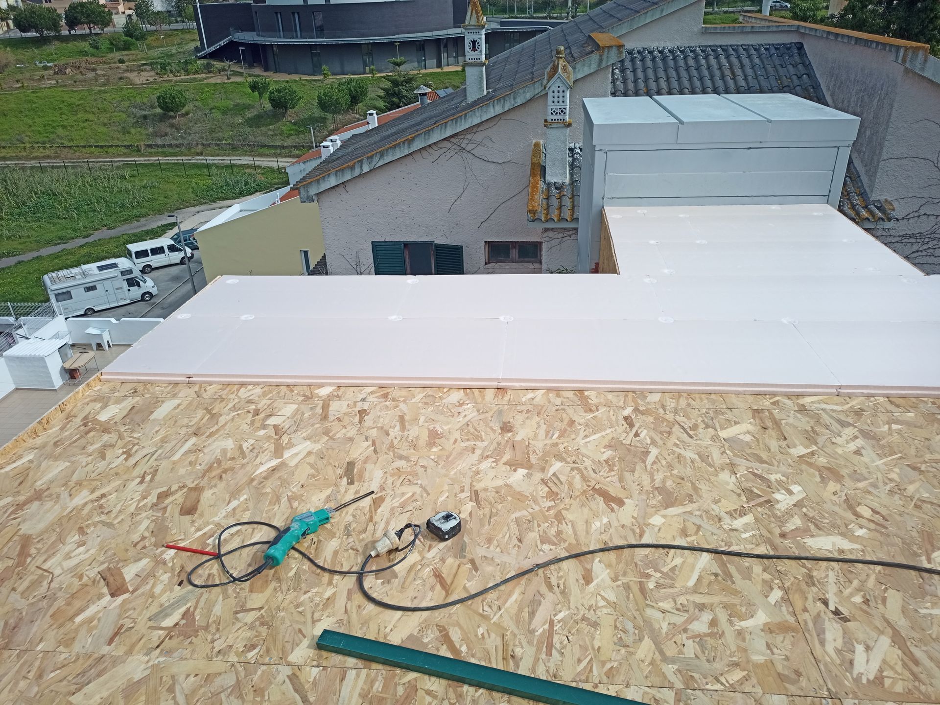 Construção no telhado: placas OSB, isolamento branco, furadeira elétrica, fita métrica. Construção em andamento, ambiente externo.