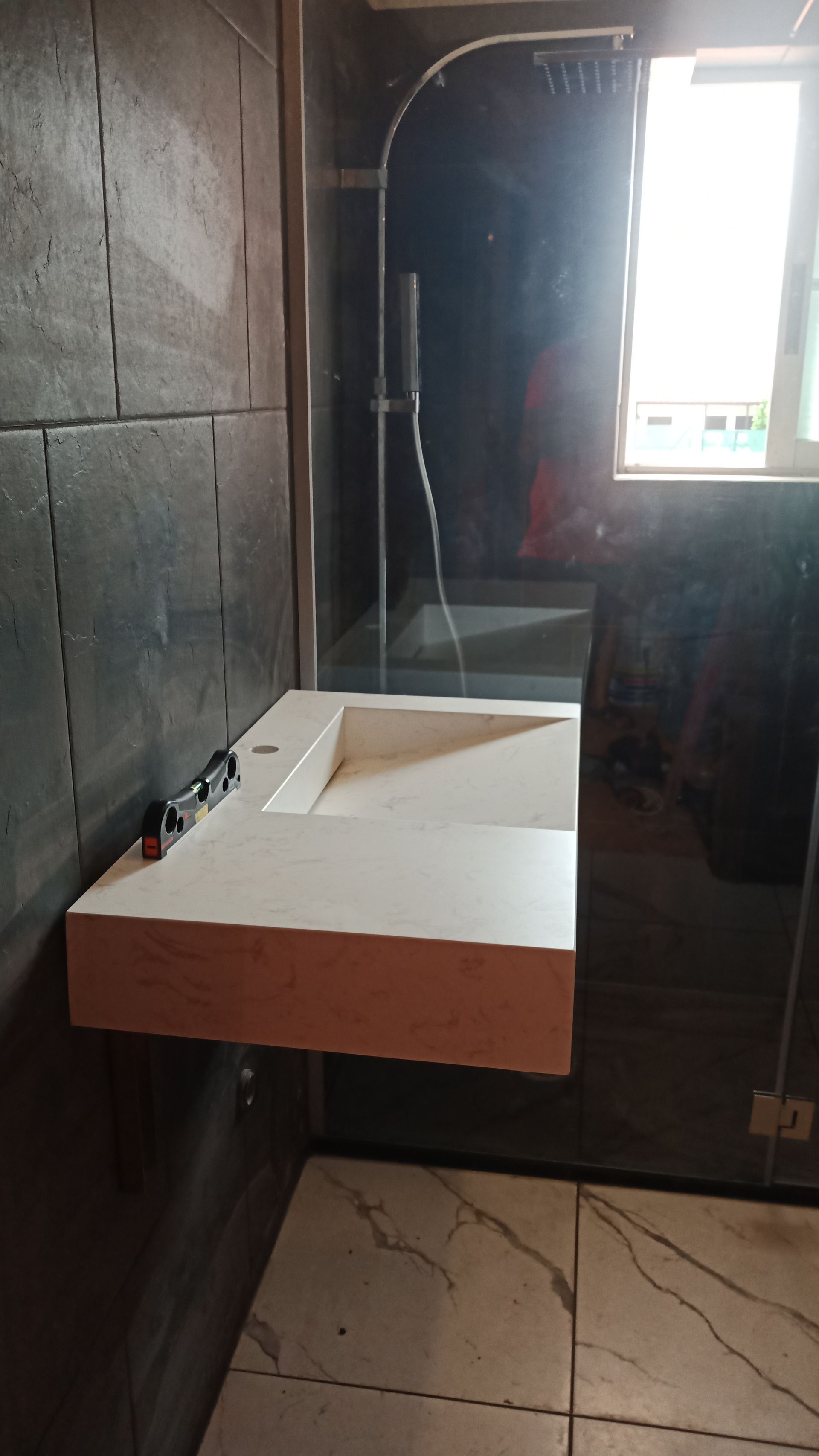 Banheiro moderno com pia branca, chuveiro, janela e paredes revestidas com azulejos cinza. Água jorrando do teto.