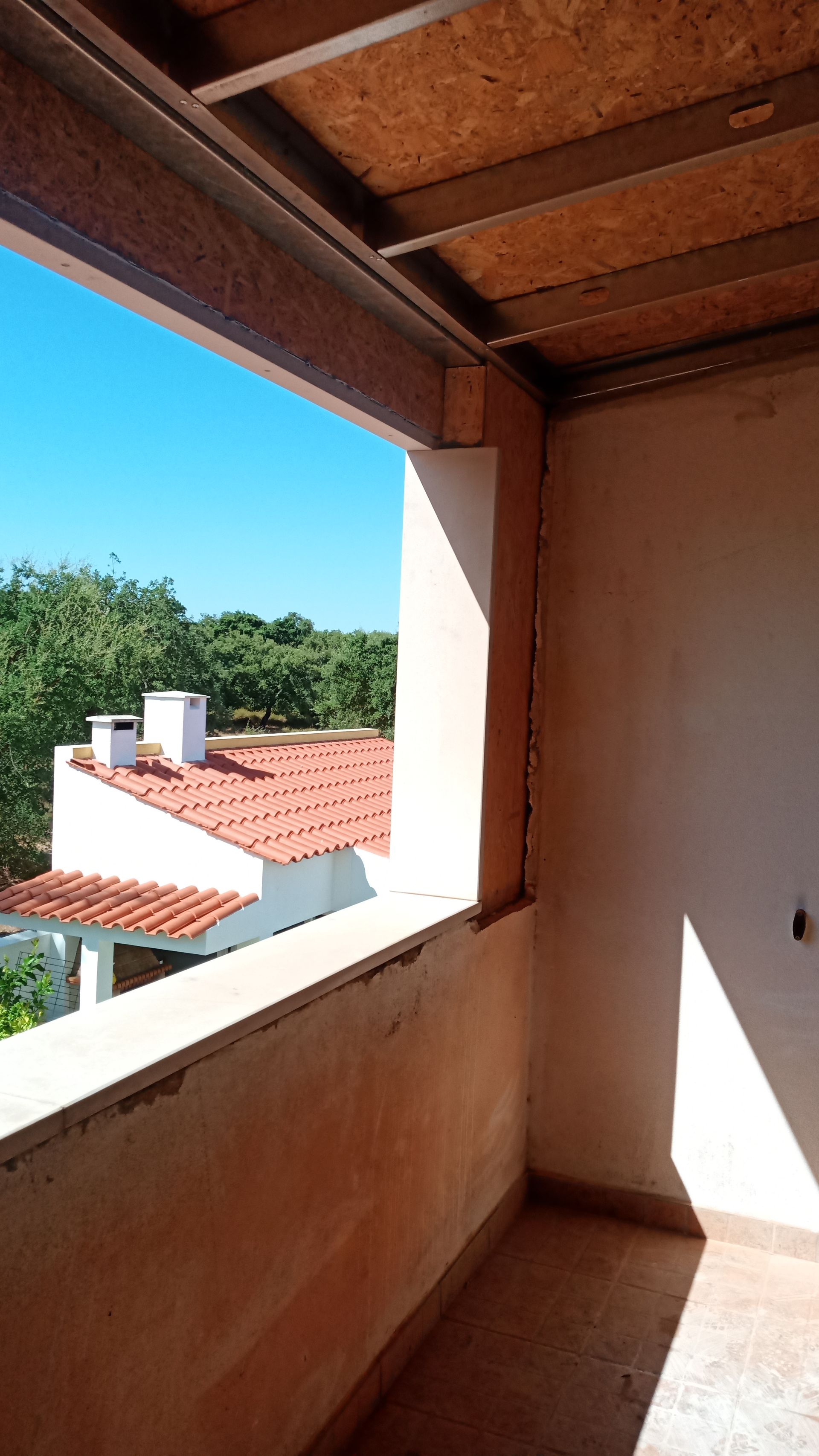 Vista da varanda: interior inacabado com estrutura aberta, com vista para telhados e árvores sob um céu azul.