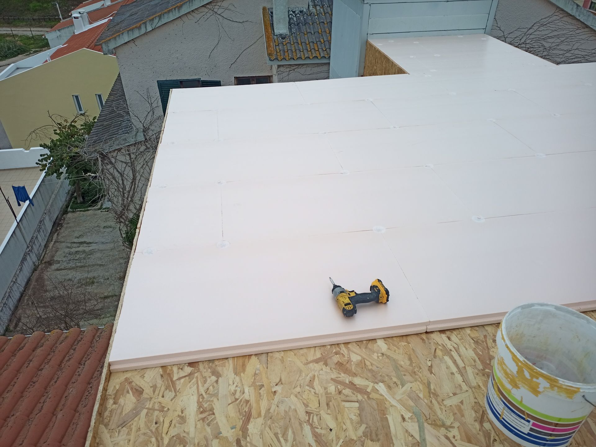 Telhado plano em construção, com painéis de isolamento rosa sobre madeira compensada. Uma furadeira e um balde são visíveis.